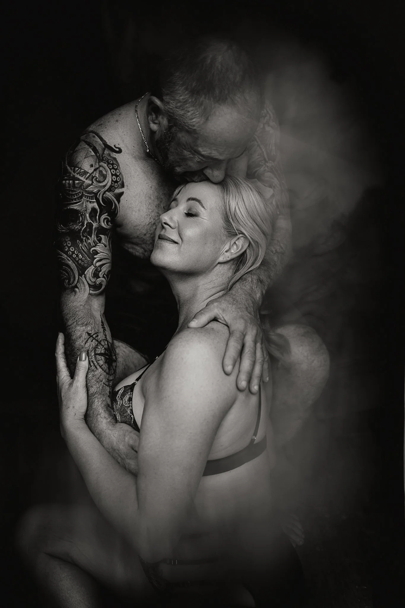 A black and white photo of an older man with tattoos and a necklace sharing a kiss with a smiling woman with light hair, wearing lingerie, in an intimate embrace.