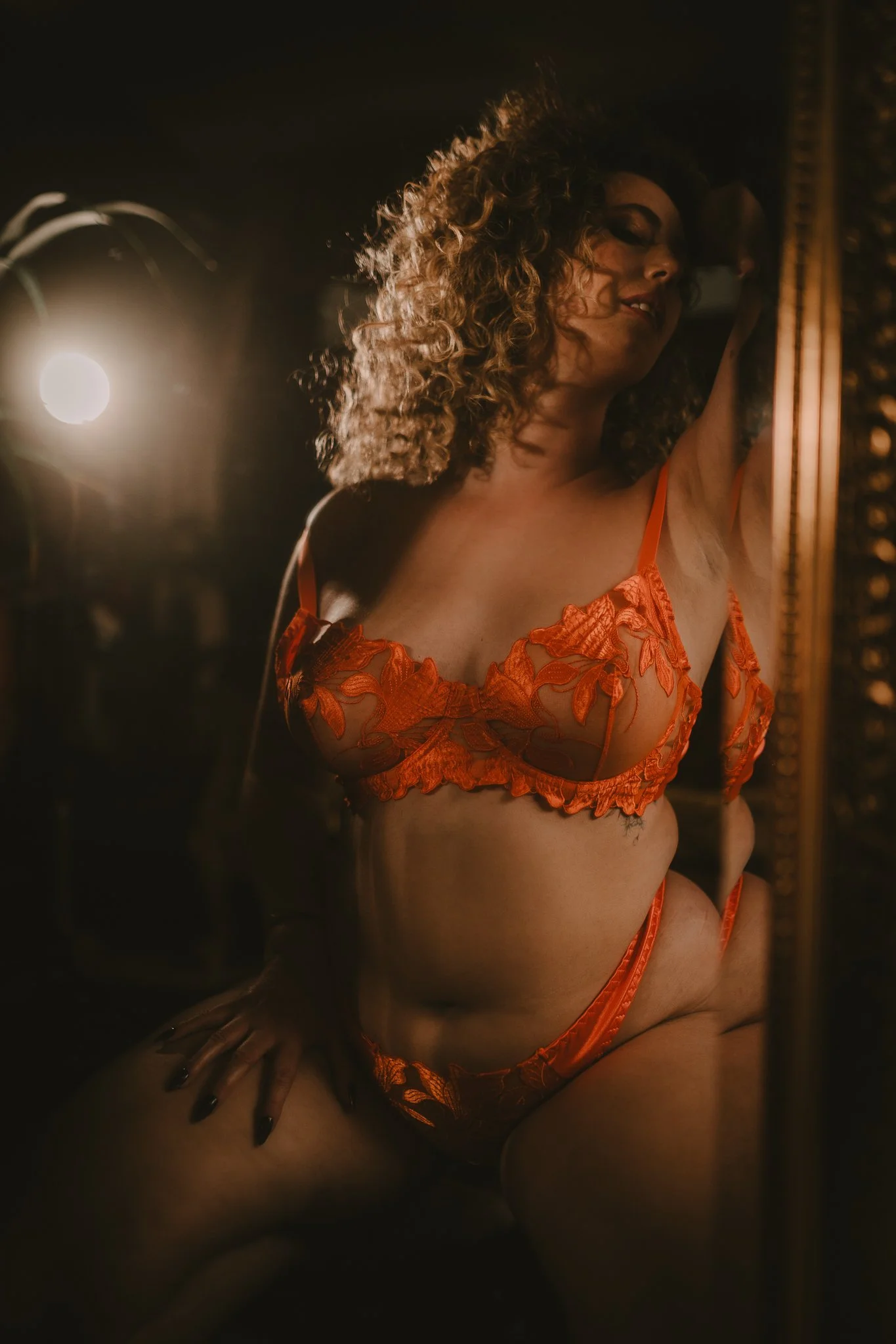 A woman with curly hair wearing orange lingerie takes a selfie in front of a mirror with dim lighting.