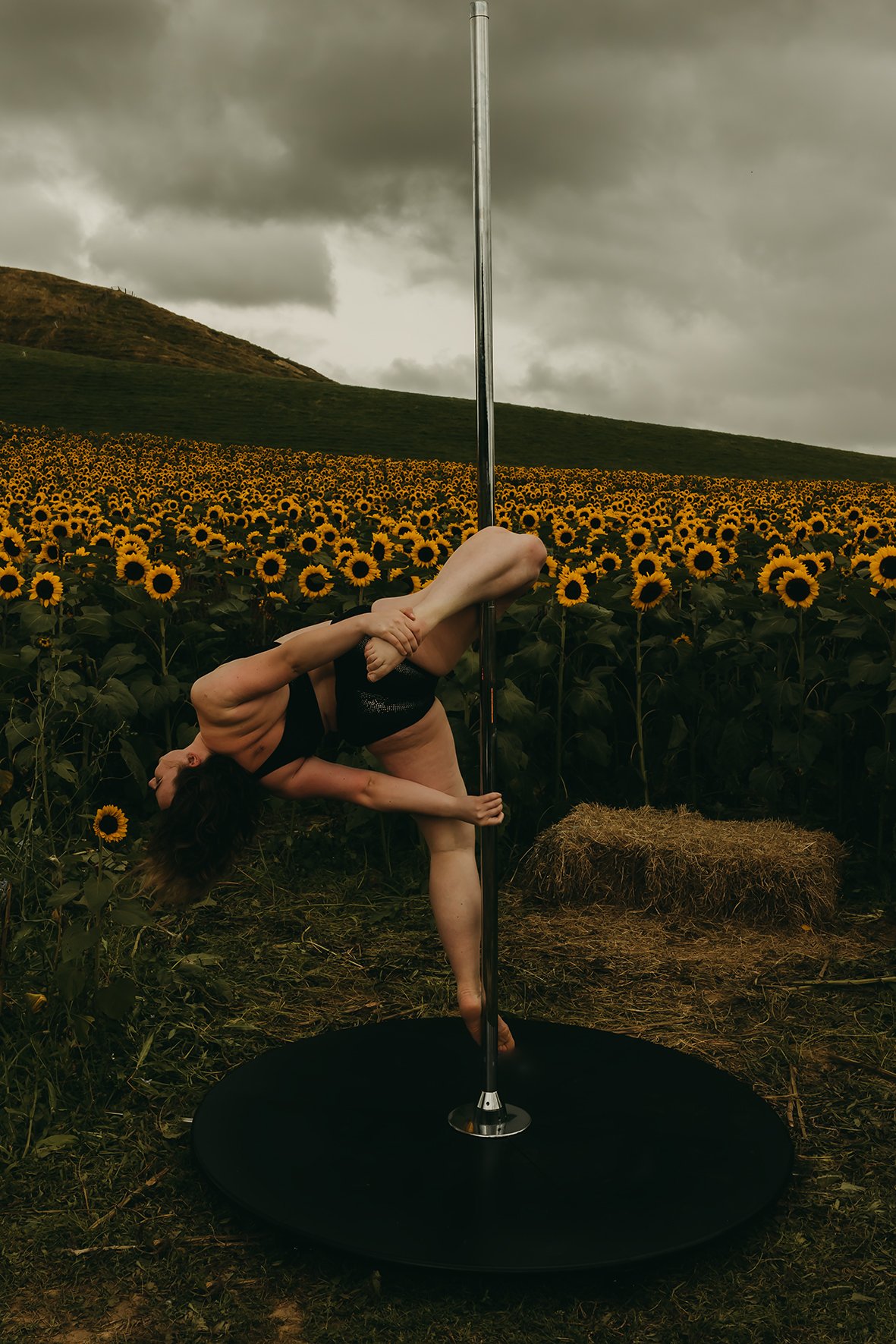 A woman performing pole dance outdoors in front of a sunflower field, with dark clouds overhead.