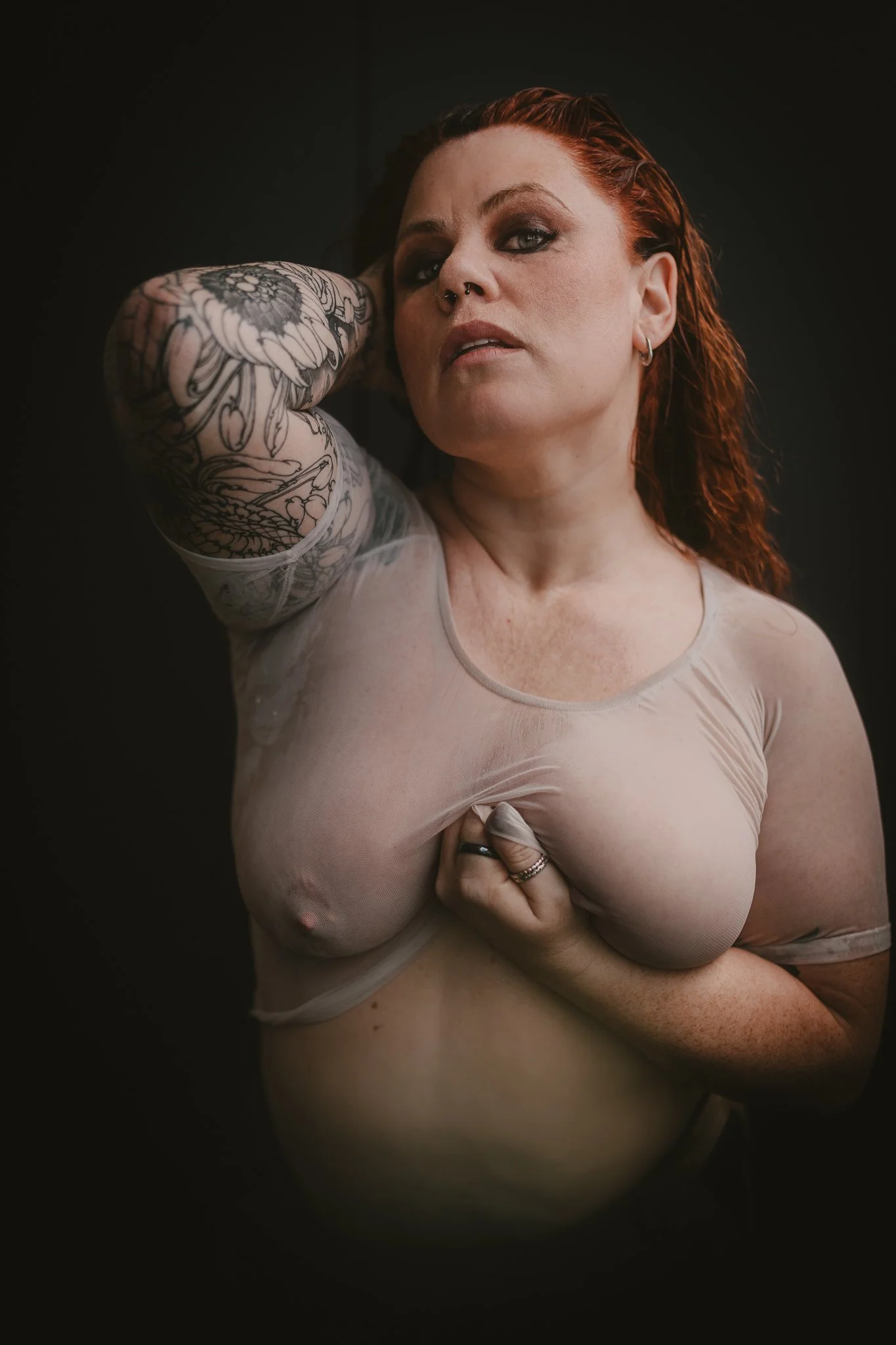 A woman with red hair and tattoos on her arm, wearing a sheer beige top, holding her breast with one hand and resting her other arm behind her head, against a dark background.