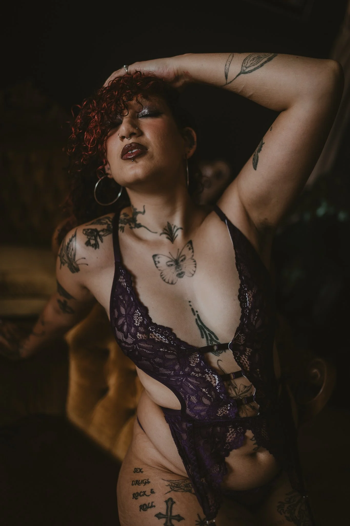 A woman with curly red hair and tattoos, wearing black lace lingerie, poses with one arm raised behind her head, showcasing tattoos and body art.
