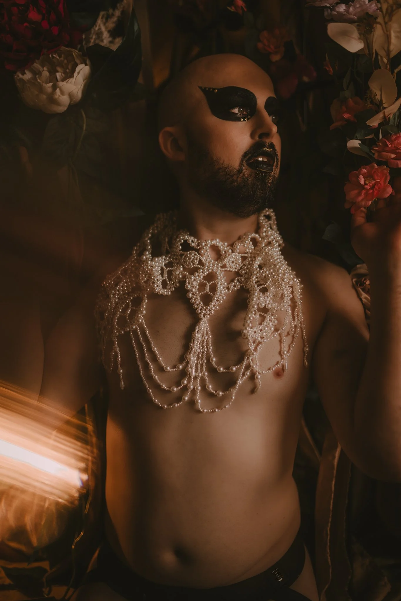 A person with shaved head and beard lying surrounded by flowers, wearing black makeup around eyes, black lipstick, and layered pearl jewelry on their chest and shoulders.