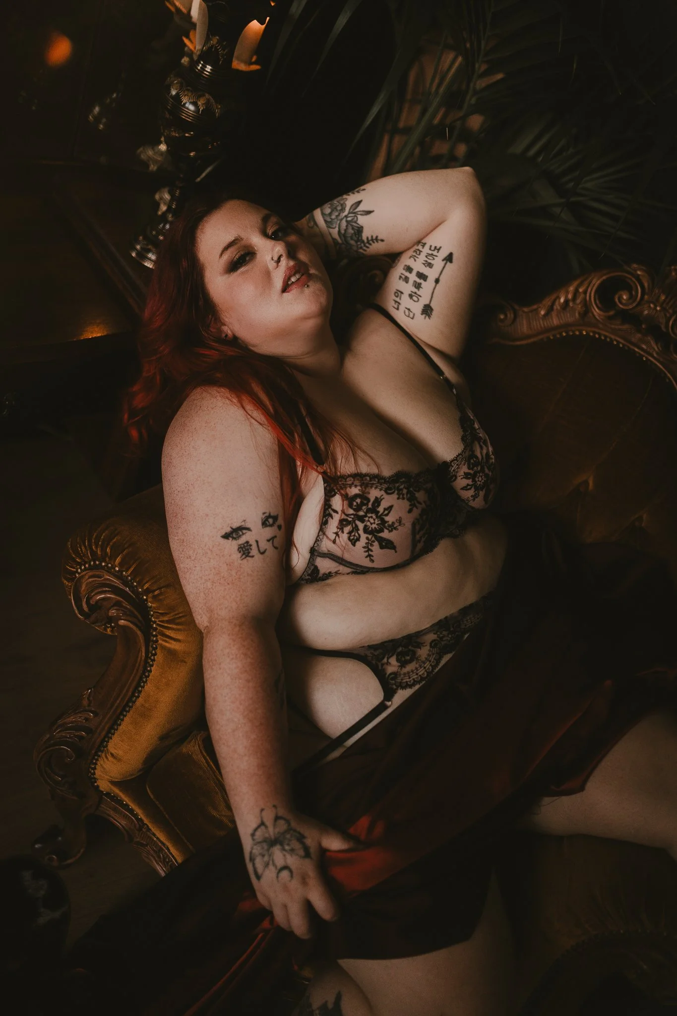 A woman with red hair, tattoos, and piercings is reclining on a vintage, ornate sofa in a dimly lit room, wearing black lace lingerie.