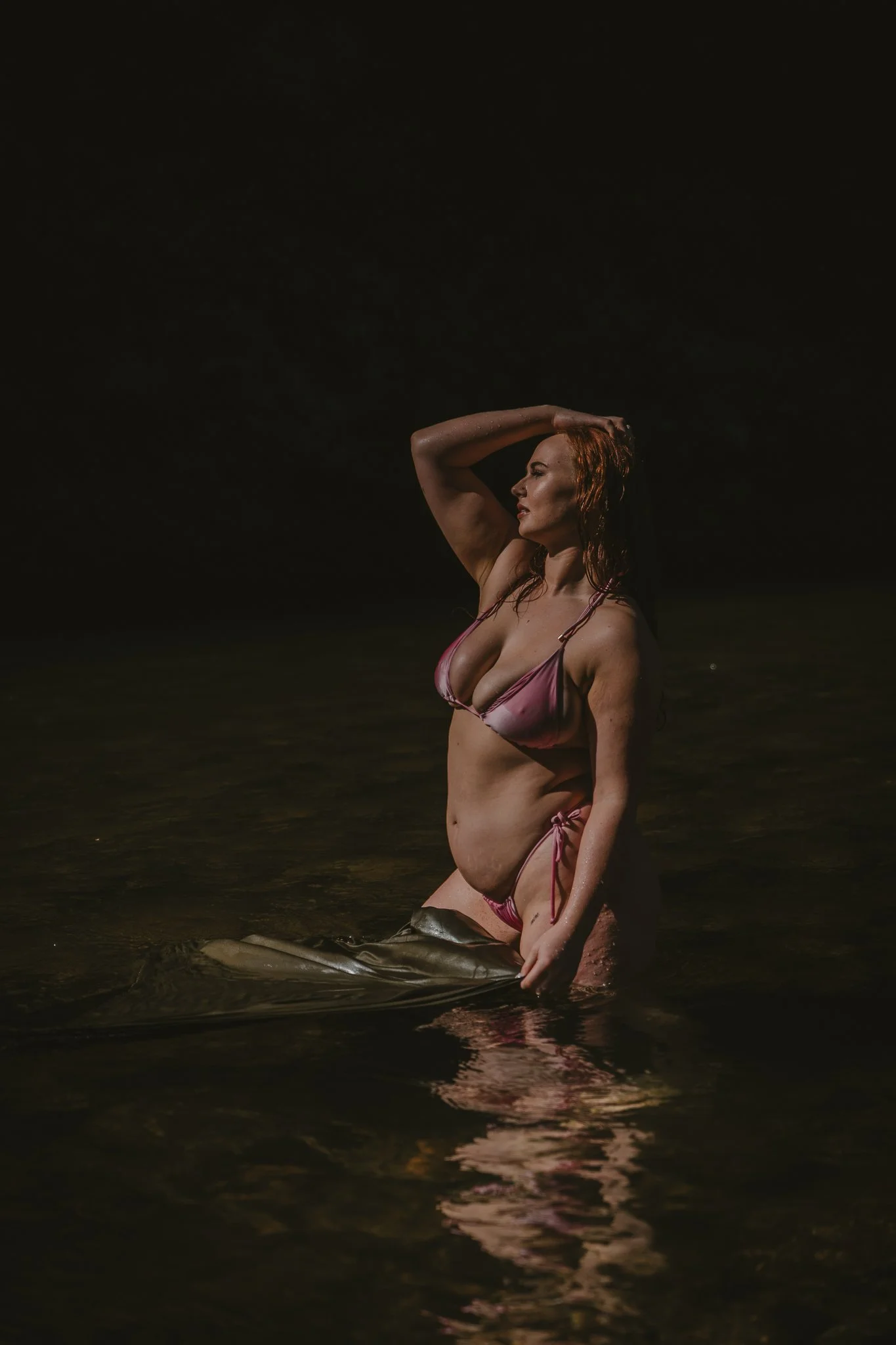 A woman in a pink bikini standing in dark water, with one arm raised and her hand resting on her head, looking to the side.