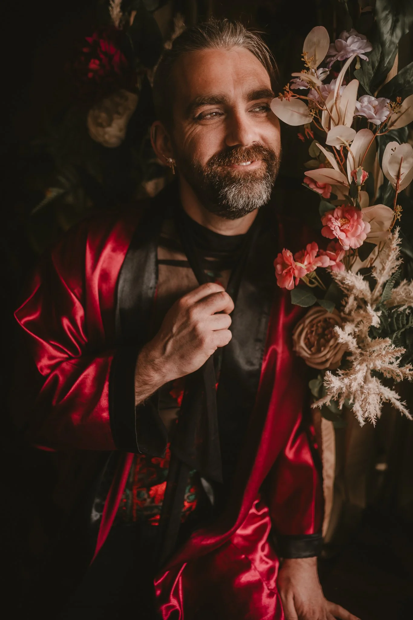 A man with a beard and long hair lying with flowers around him, wearing a shiny red jacket and black clothing, smiling softly.