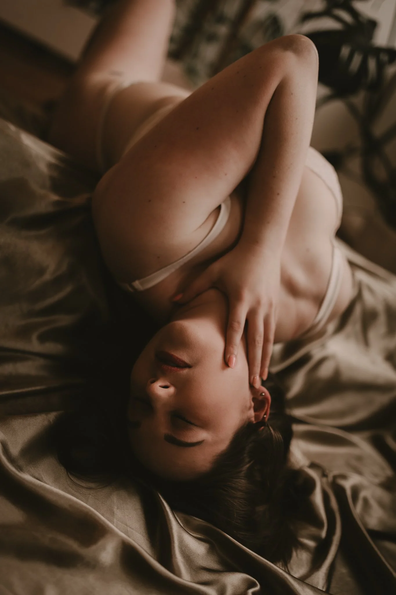 A woman lying on her back on a bed, wearing a beige bra, with one hand on her face and her eyes closed.