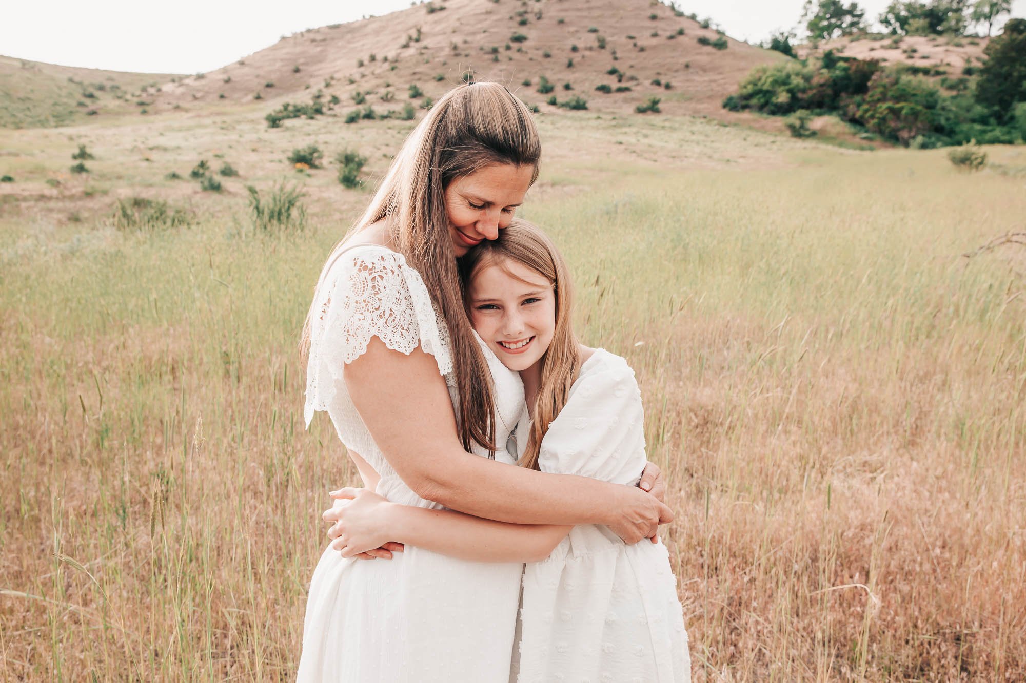 Woman and girl in white dresses hugging in a green meadow with hills in the background.
