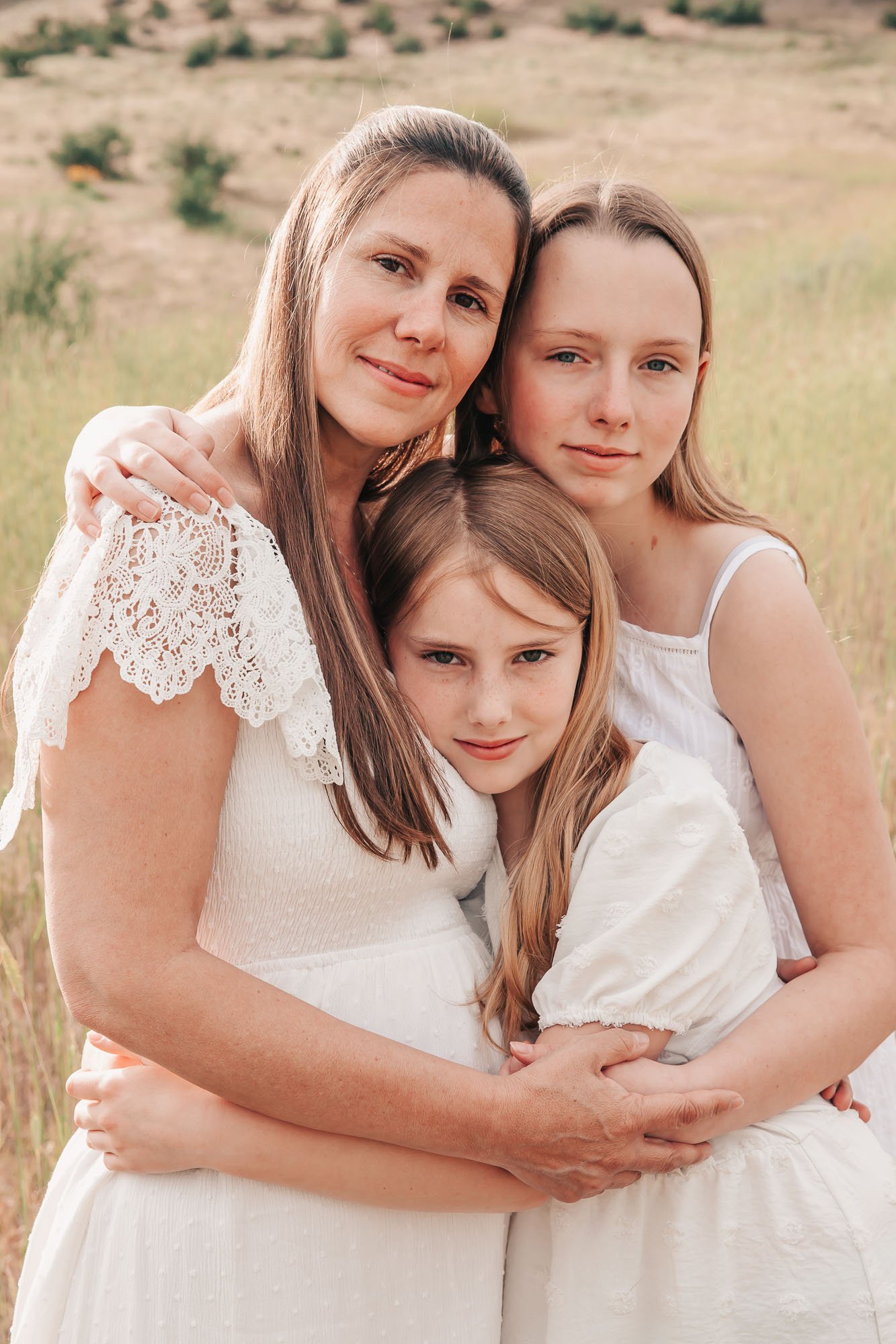 Mother and two daughters in white dresses embracing outdoors in a field.