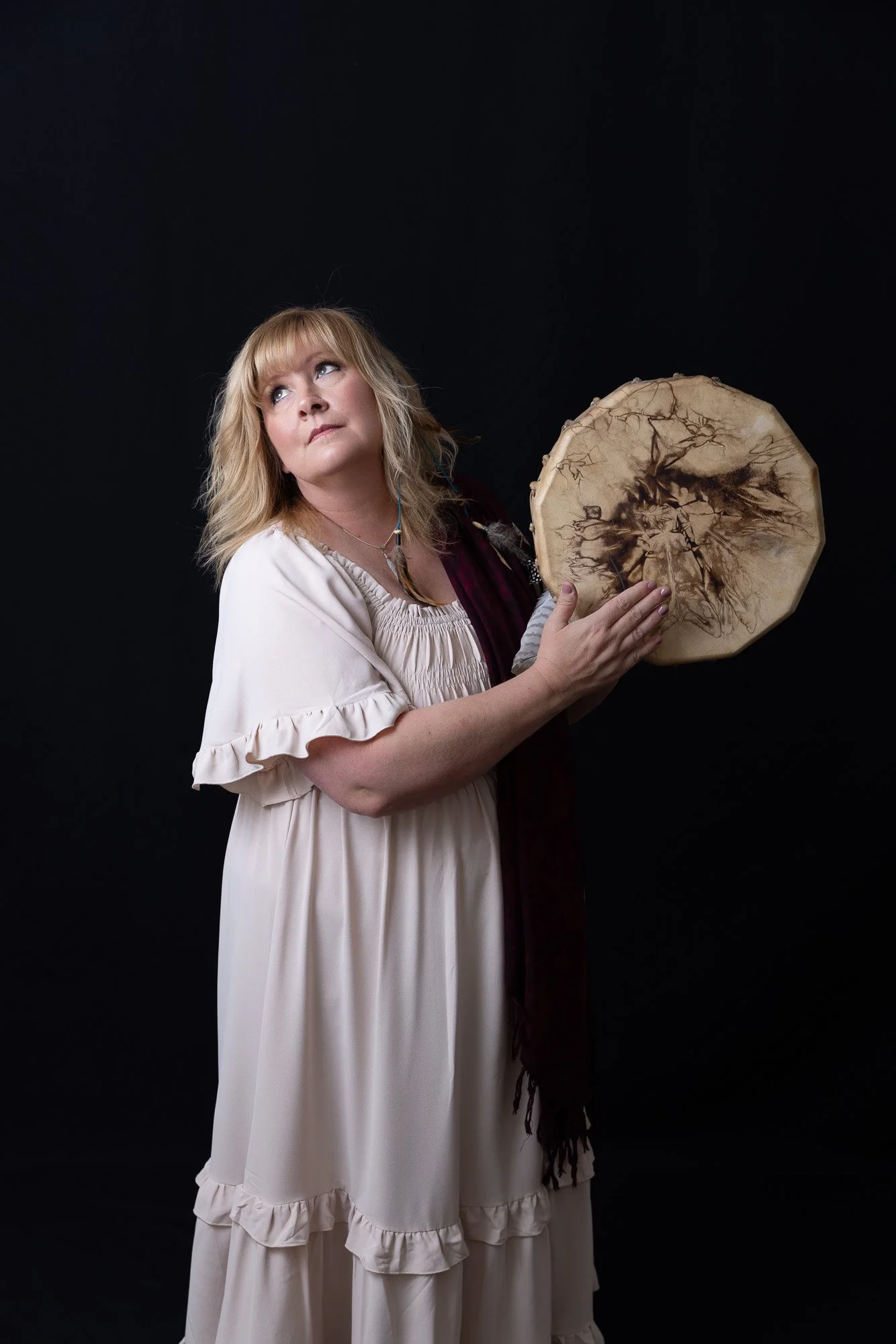 Portrait of a shamanic medicine woman playing a drum. She is wearing a cream colored dress and gazing up and over her shoulder.
