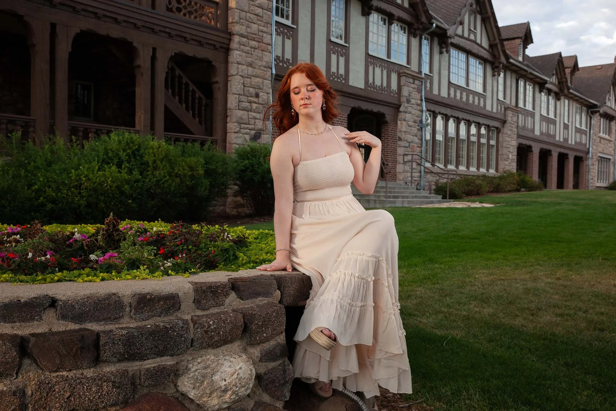 A woman with red hair in a cream-colored dress sitting on a stone wall near a garden with colorful flowers, with a large stone and wood house in the background.
