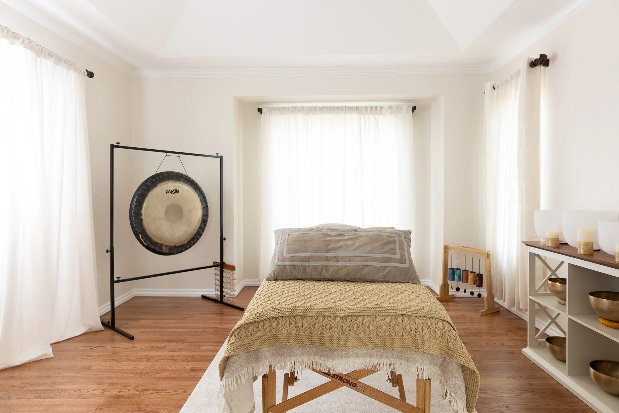 Photo of a sacred sound healing meditation space. The room is bright with natural light and lots of windows, a massage table in the middle, a gong in one corner, chimes in another corner, and a cubby shelf full of Tibetan singing bowls.