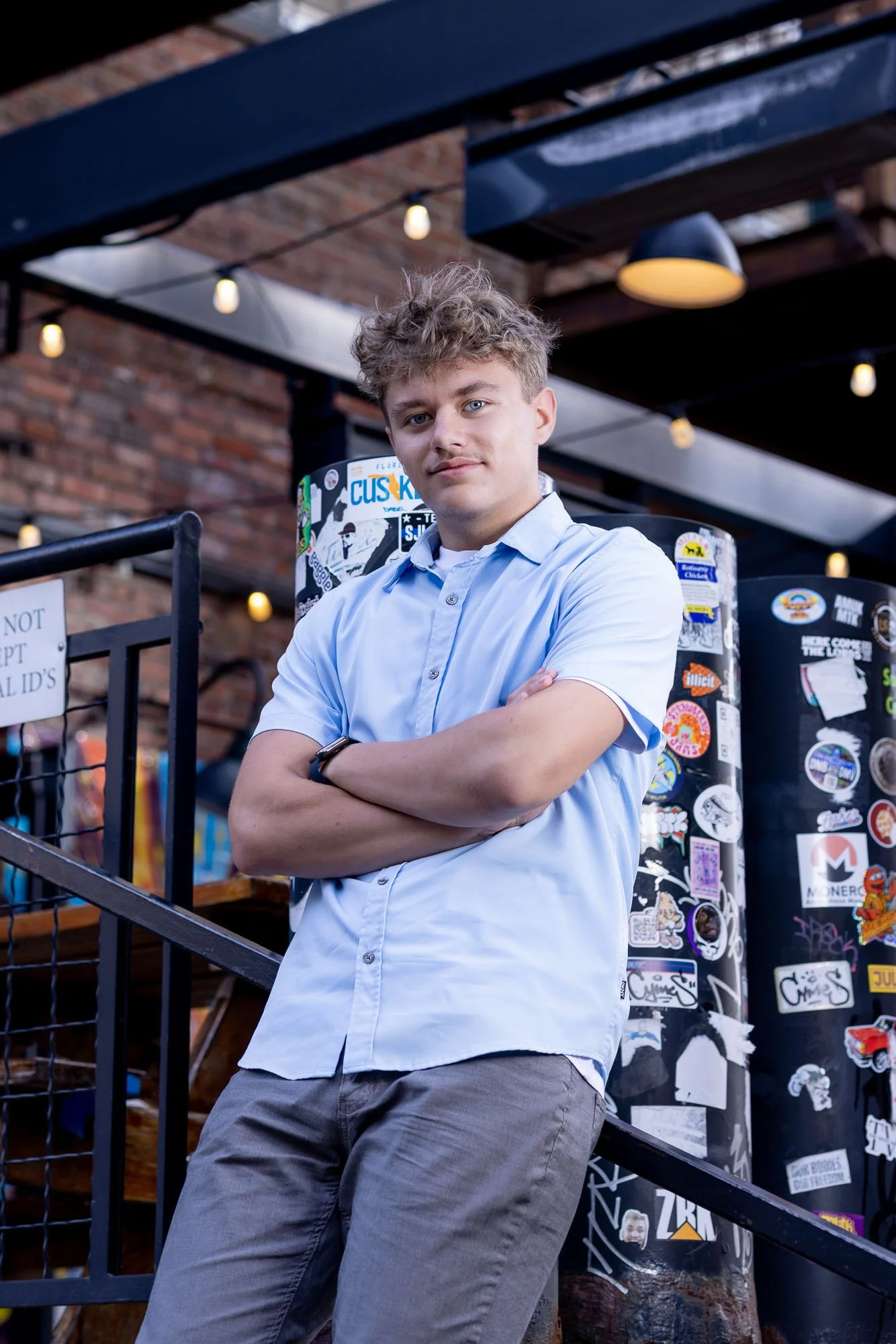 Senior portrait of a young man with his arms crossed, looking straight into the camera. String lights from a patio bar are lit up behind him.