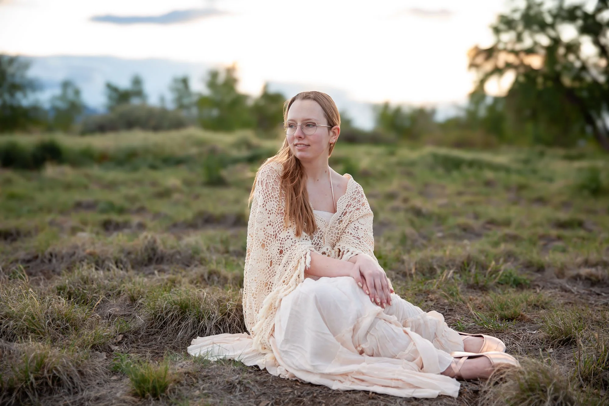 A woman with long red hair and glasses sitting on the ground in a grassy field, wearing a cream-colored lace shawl and dress, with trees and a cloudy sky in the background.