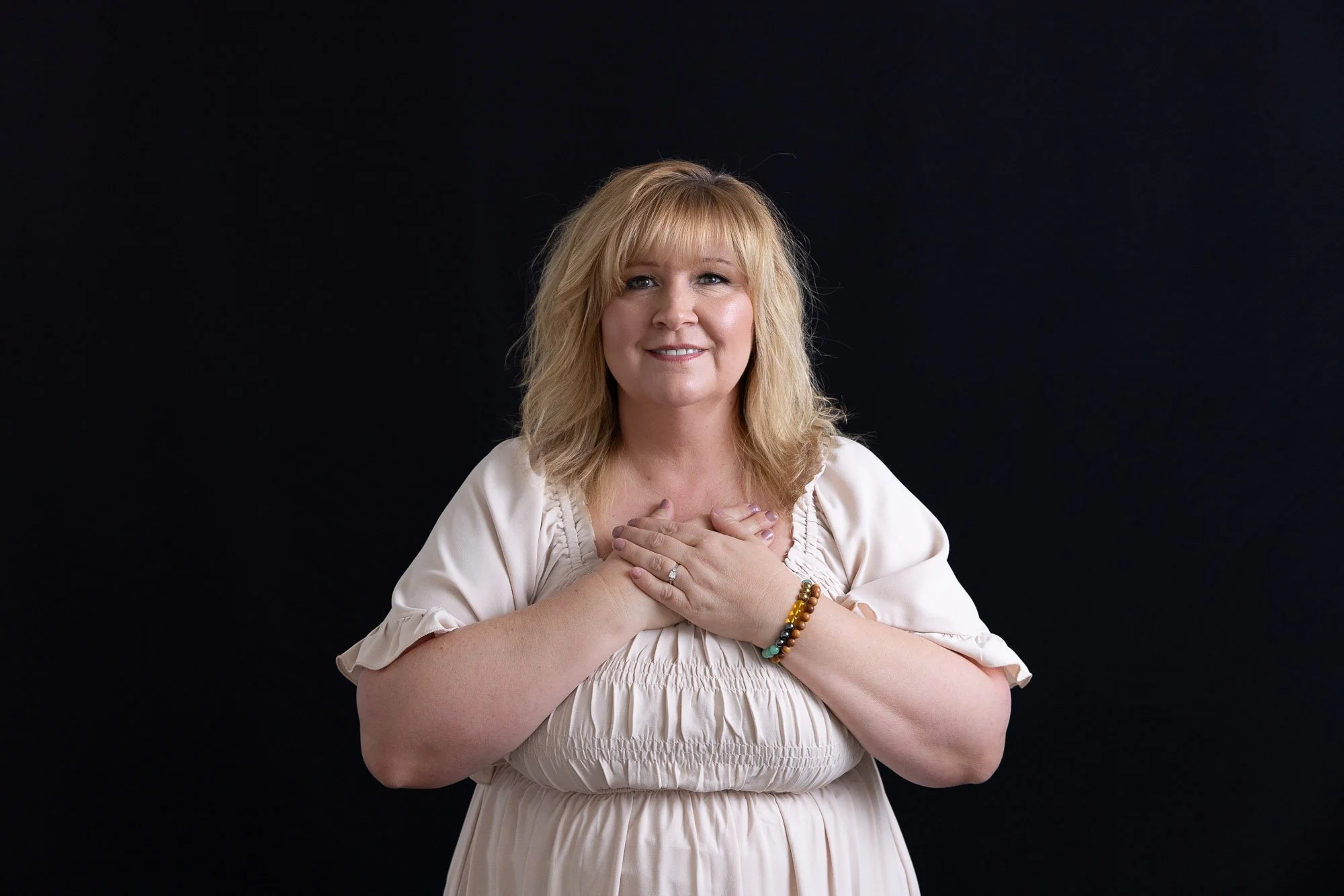Portrait of a blond woman in a cream colored dress, set against a black backdrop. She is holding her hands over her heart, gazing straight at the camera, and smiling.