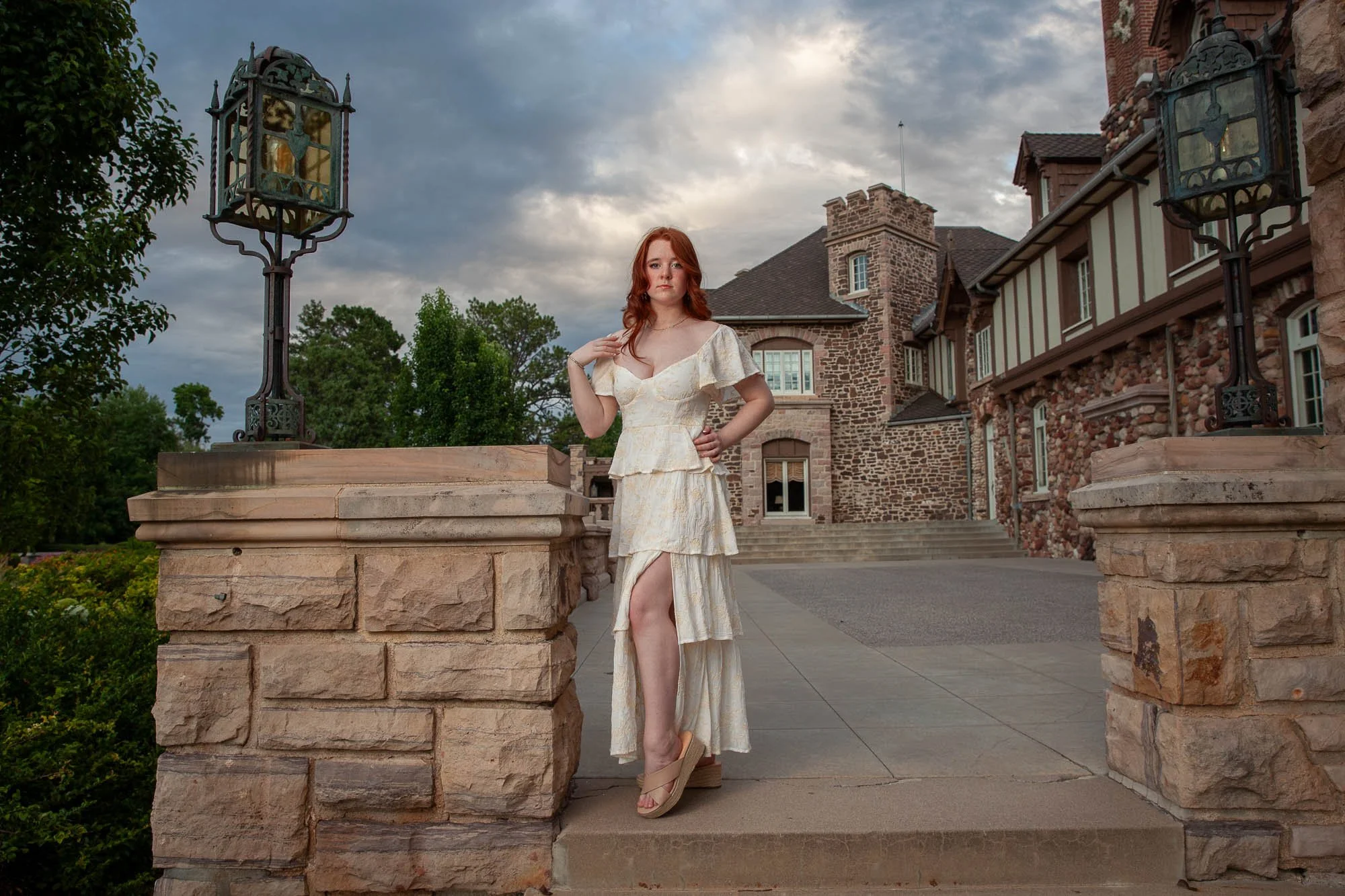 A woman in a cream-colored, layered dress with a high slit stands on the steps of a large stone and brick mansion, posing with one hand on her hip and the other near her shoulder. The mansion has multiple chimneys, boarded-up windows, and decorative