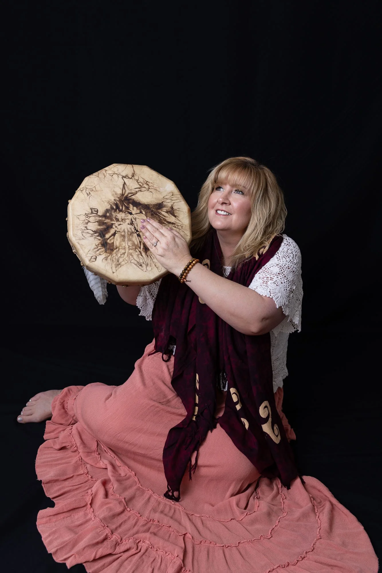 Portrait of a shamanic medicine woman holding a drum. She is dressed in a cream colored lace knit top with a pink ruffle skirt and a wine colored scarf.