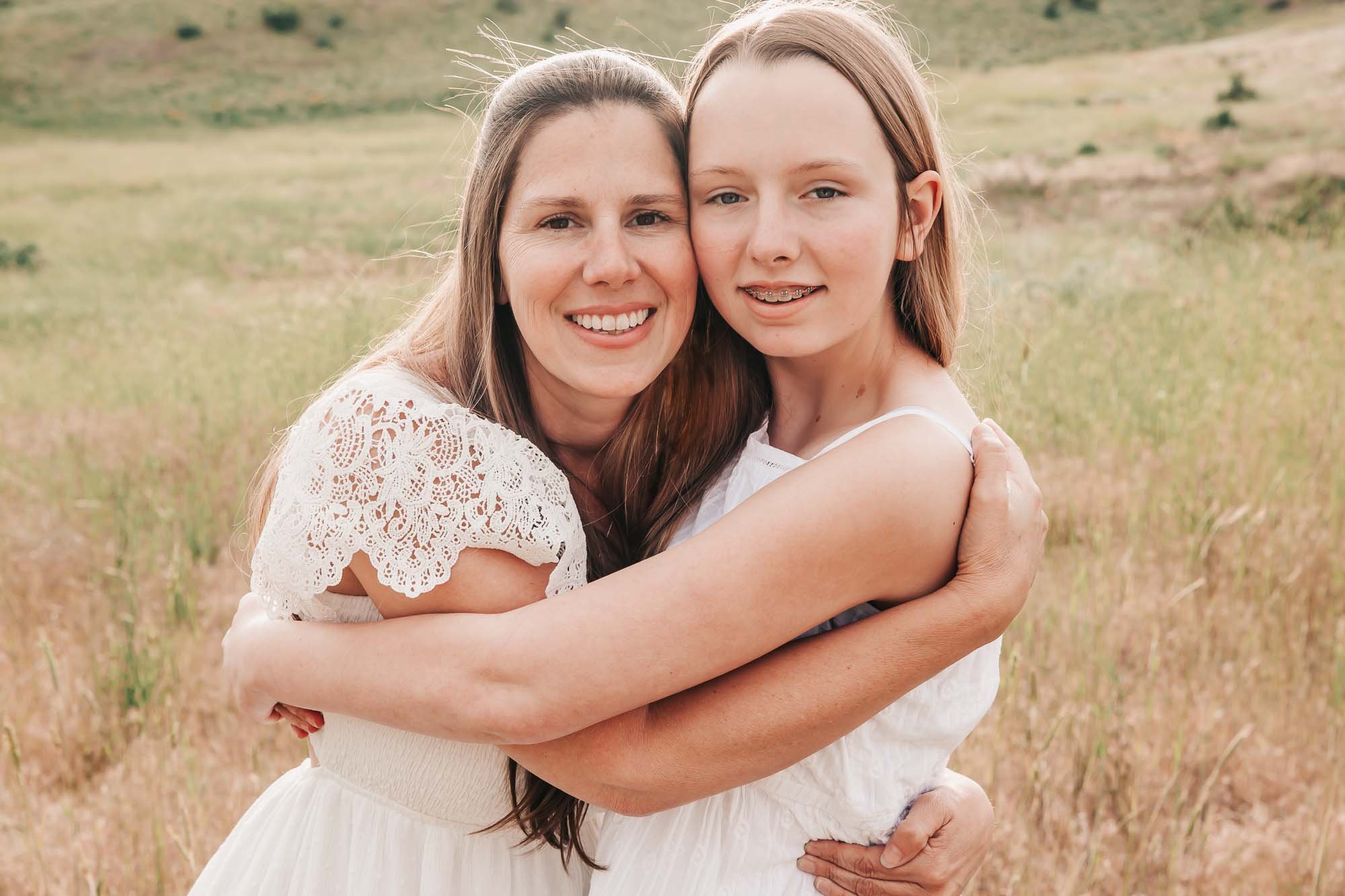 Mother and daughter hugging in a grassy field; both wearing white dresses.