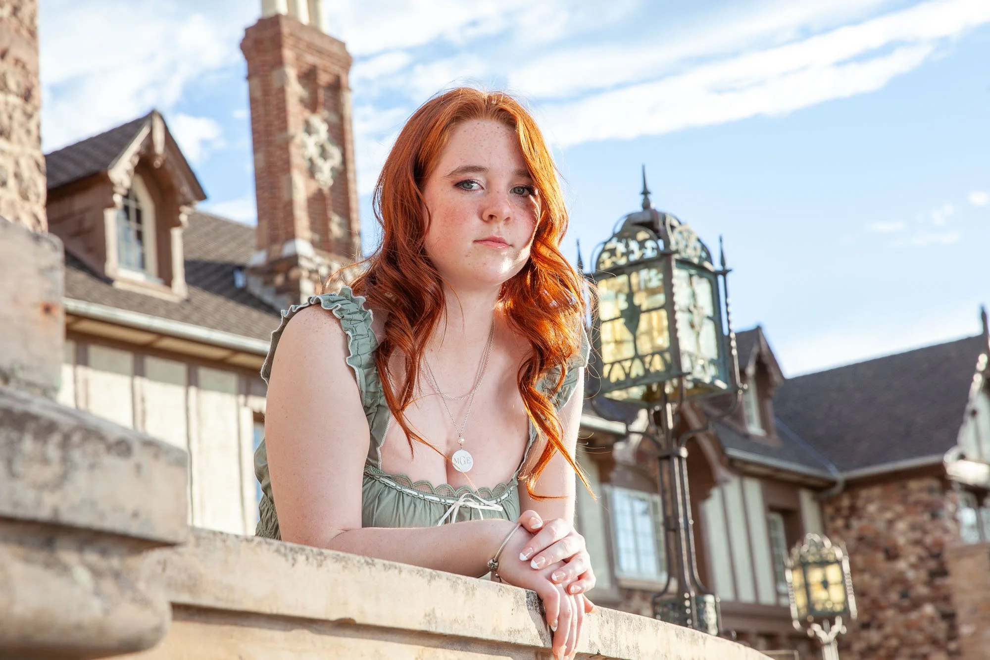 Young woman with red hair leaning on a stone ledge outdoors with historic buildings and a lamp post in the background.