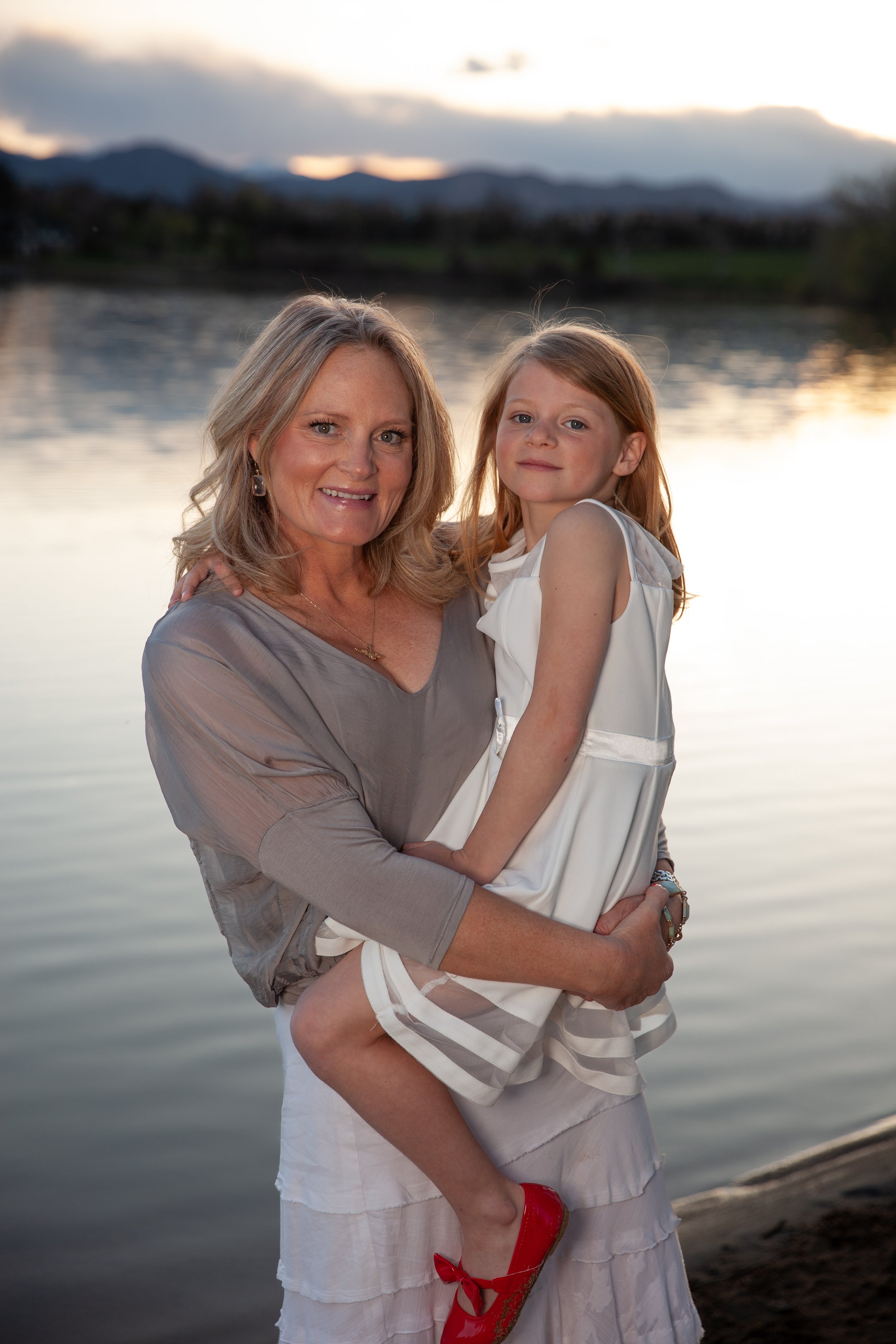 A woman holding a young girl near a lake at sunset, with mountains in the background. The woman is wearing a light gray top and white skirt, and the girl is in a white dress with red shoes. The scene is serene with a water reflection and soft lightin