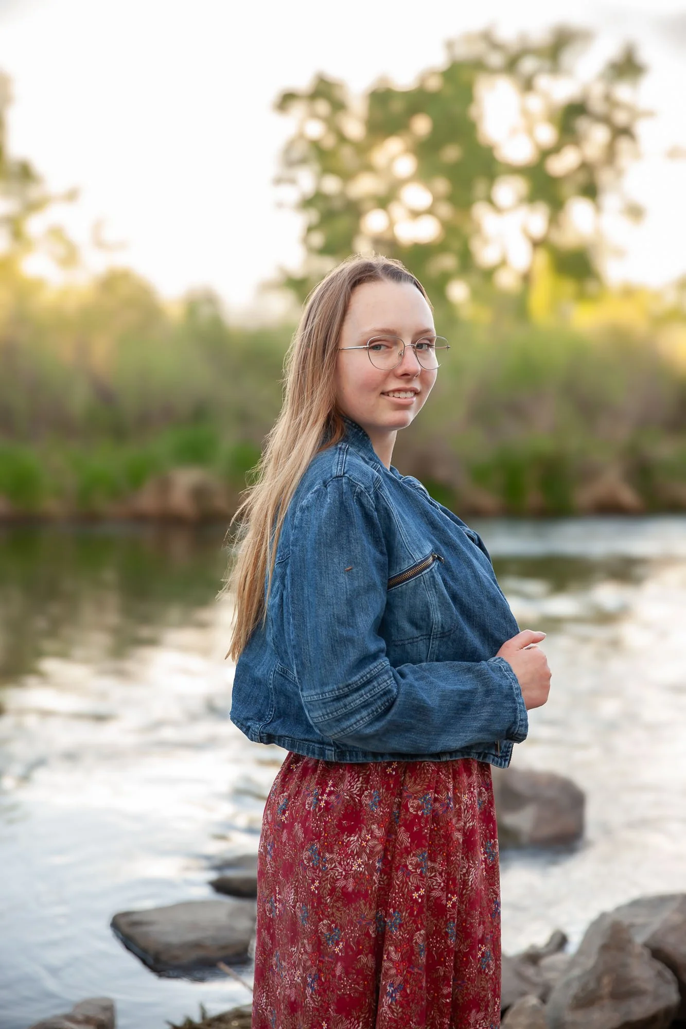 A young woman with long, blonde hair and glasses stands by a river at sunset, wearing a denim jacket and a long, red patterned skirt.