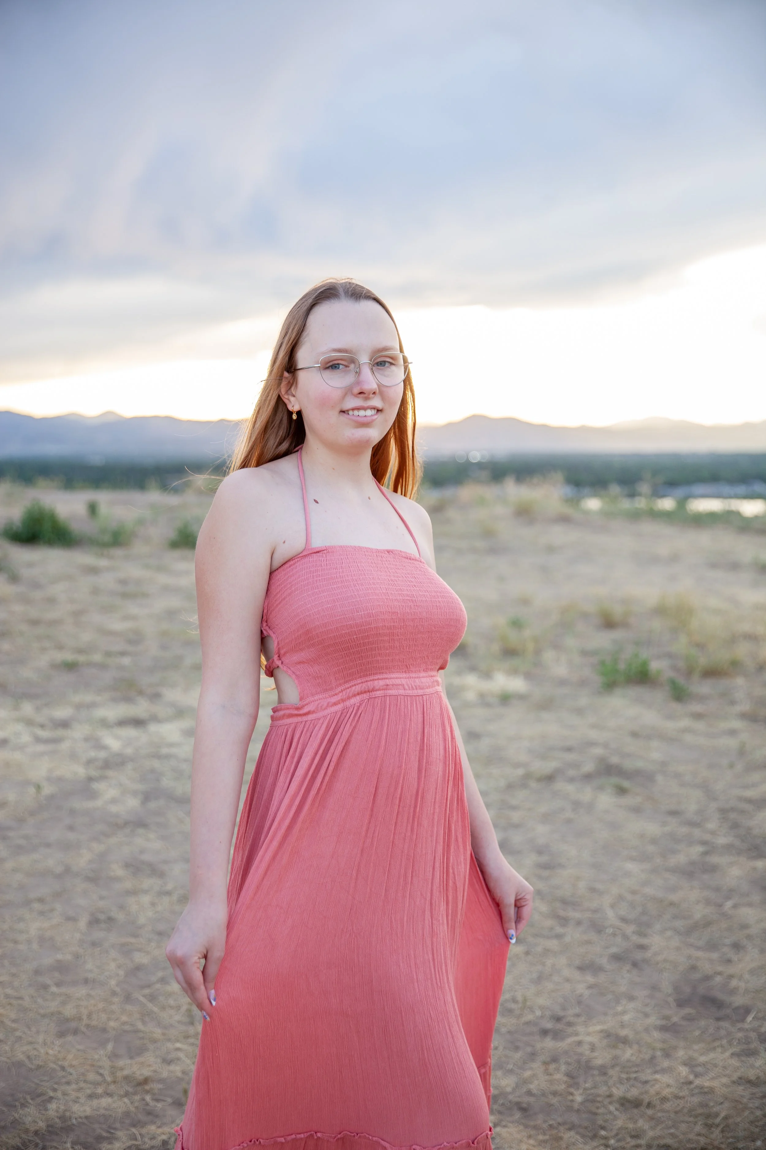 A young woman with glasses and long blonde hair standing outdoors in a natural landscape during sunset, wearing a coral pink sleeveless dress.