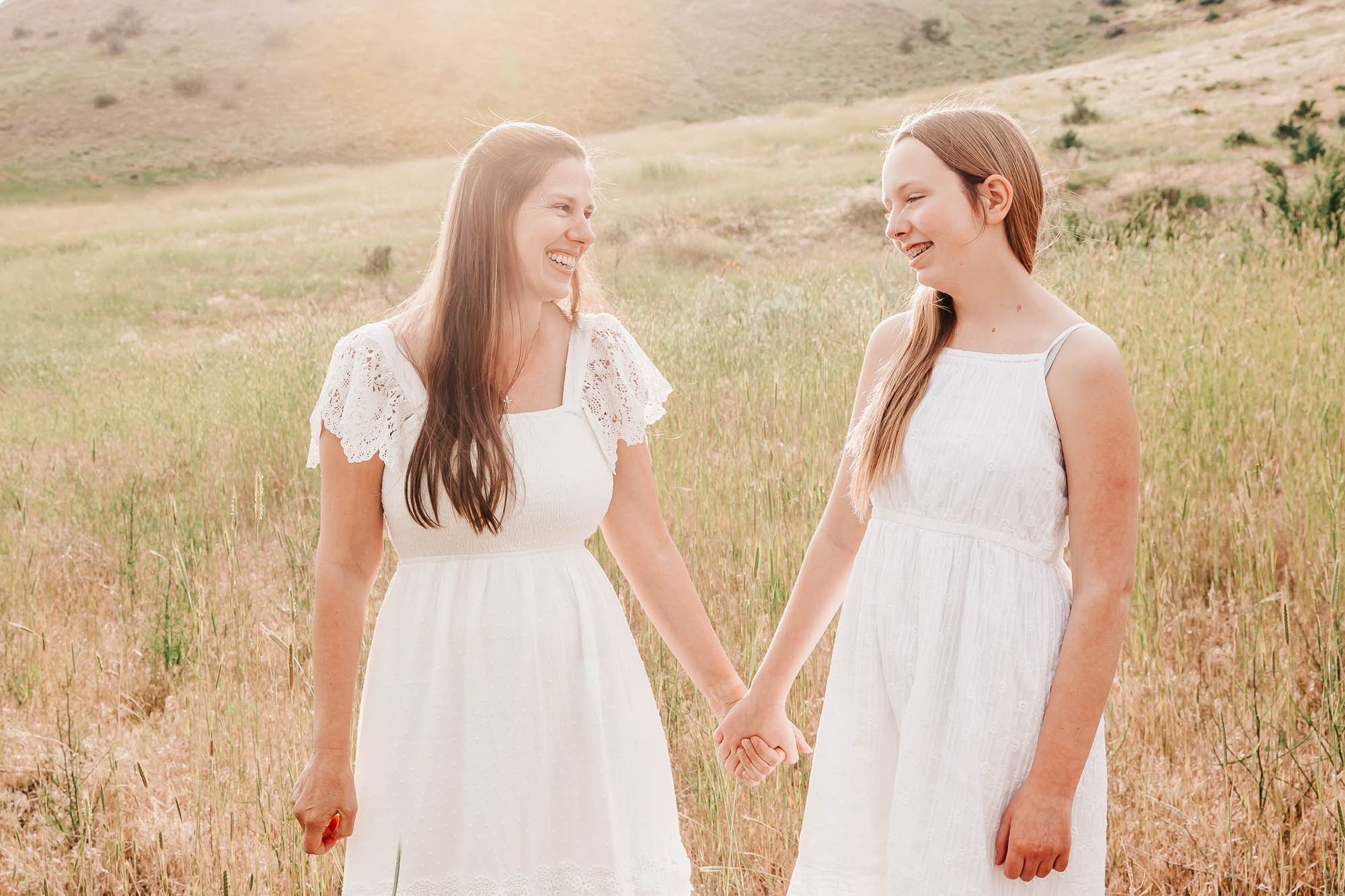 Two women in white dresses holding hands and smiling in a grassy field with sunlight in the background.