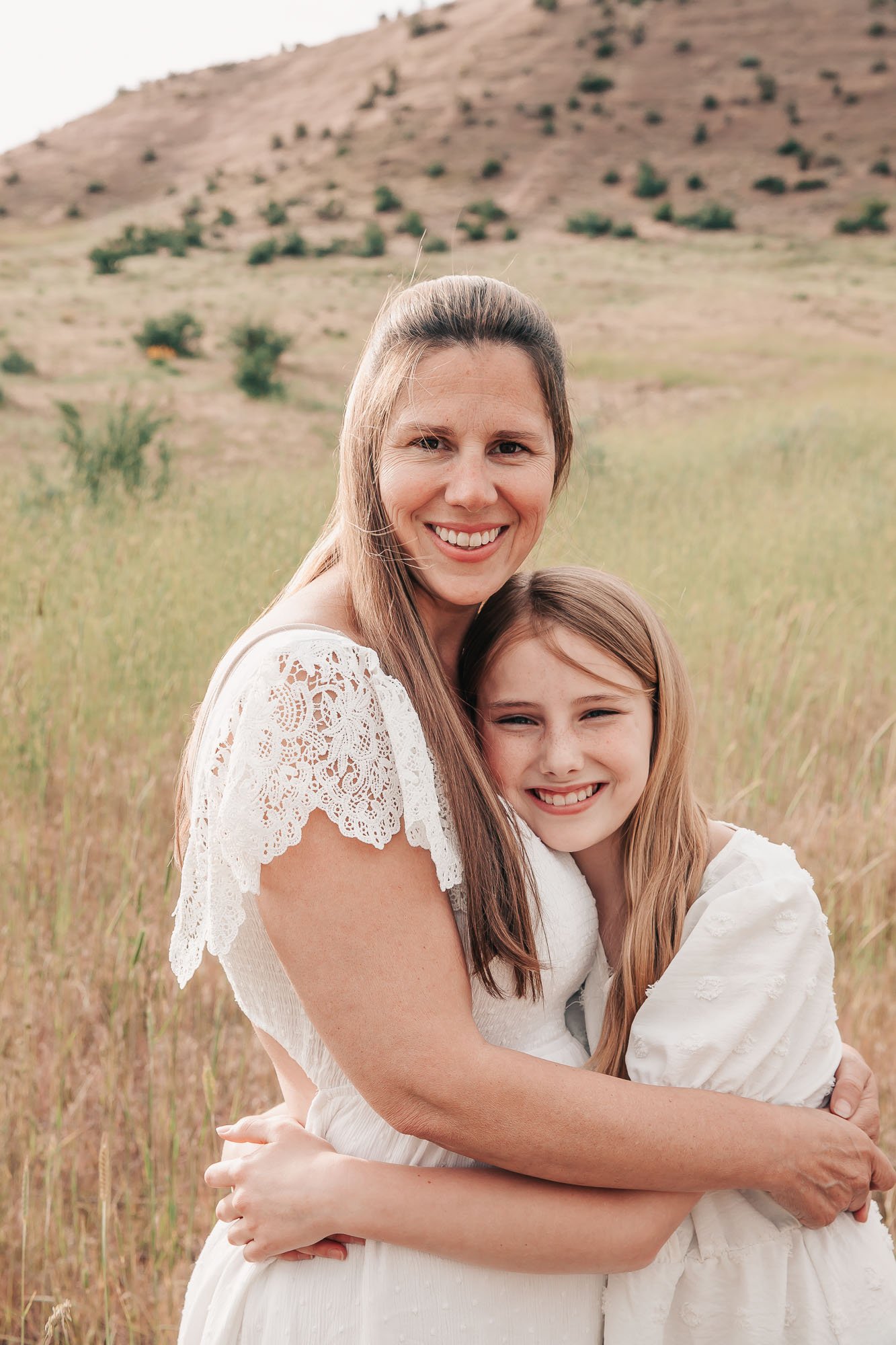 A woman and a young girl hugging and smiling in a grassy field with hills in the background.