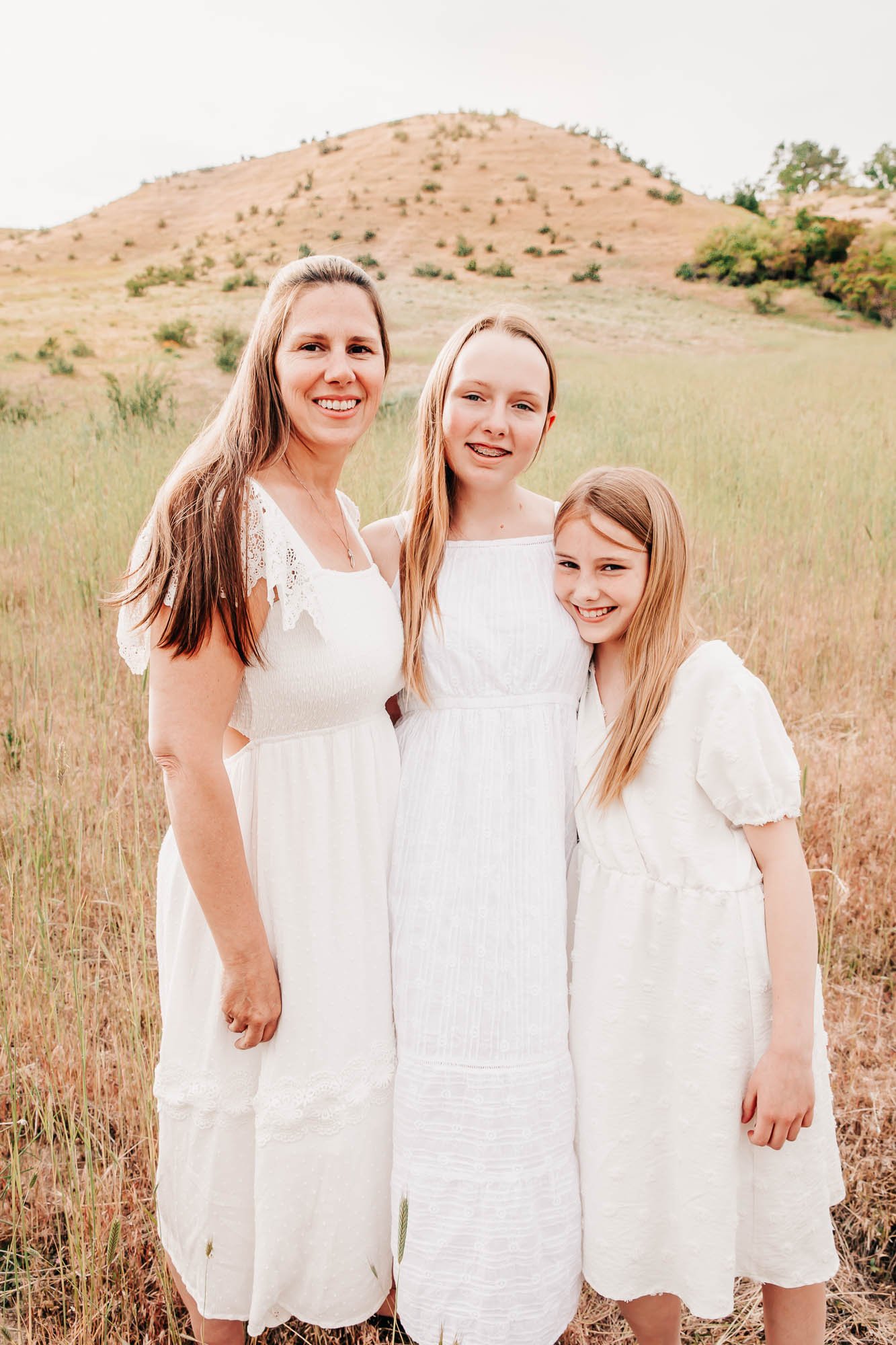 Three females in white dresses standing in a grassy field with a hill in the background.