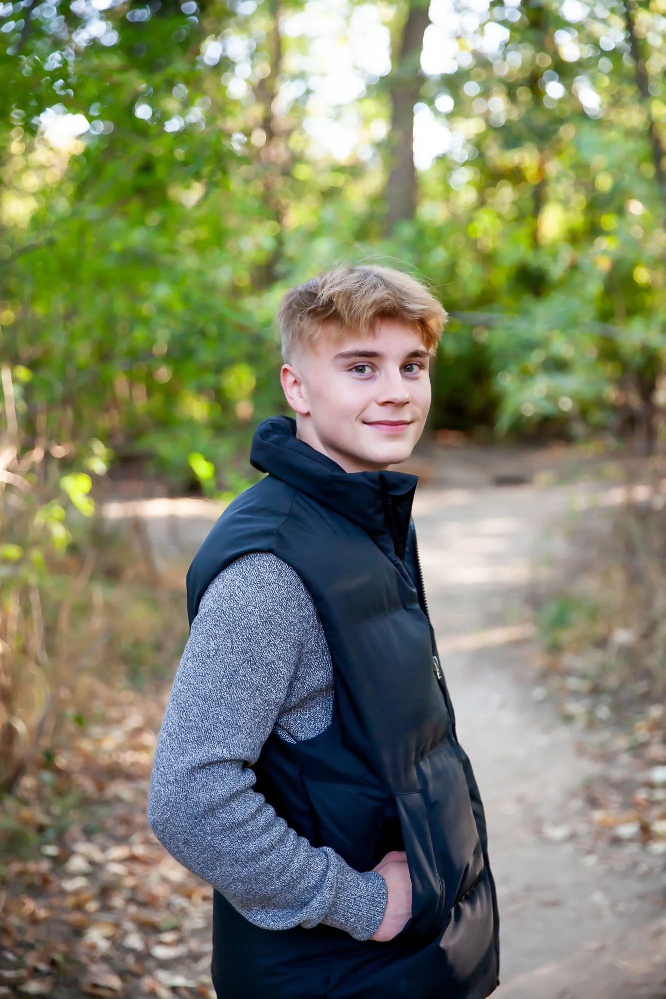 A young man with blonde hair, wearing a grey sweater under a black vest, stands on a wooded trail with trees and greenery in the background, smiling at the camera.