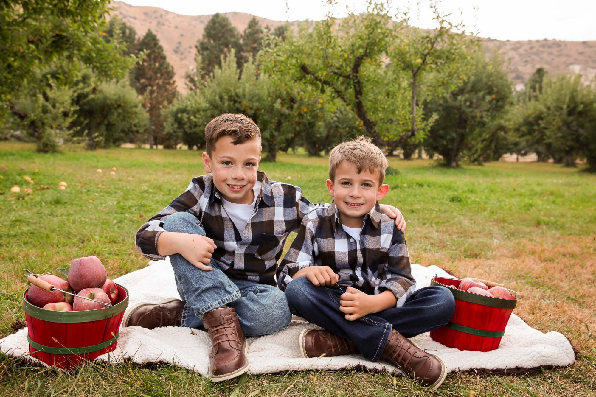 Two boys sitting on a blanket in an orchard with apples in buckets.