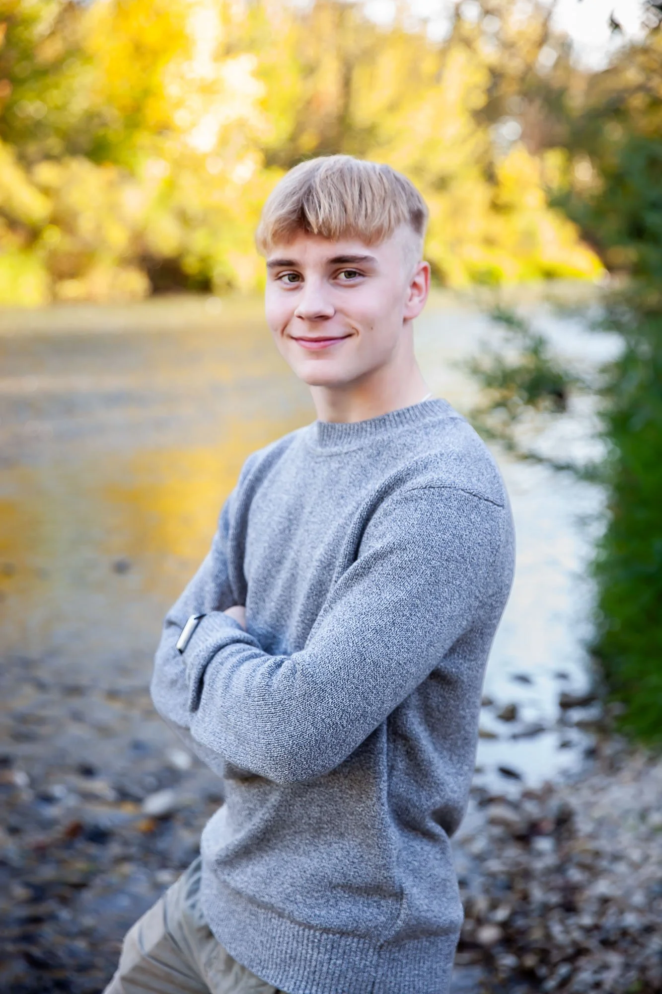 A young man in a gray sweater with crossed arms smiling outdoors near a river, with trees showing autumn foliage in the background.