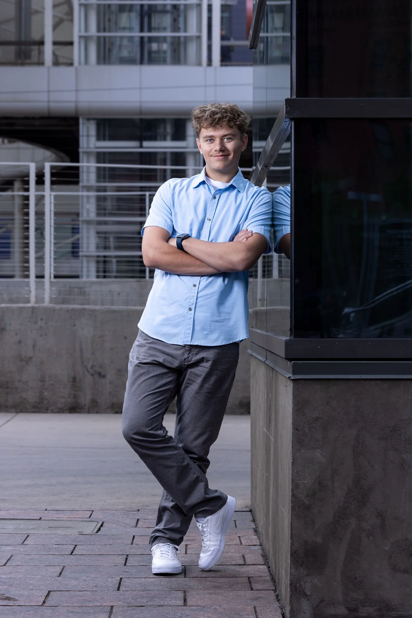 Senior portrait of a young man leaning agains a reflective wall in an urban setting. He has a blue button down shirt and is crossing his arms and smiling at the camera.