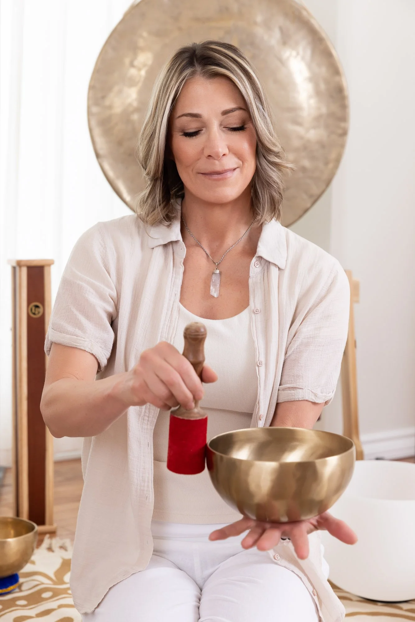 Portrait of a woman, a sound healer, playing a Tibetan singing bowl. There is a gong behind her and she has a beautiful expression on her face.