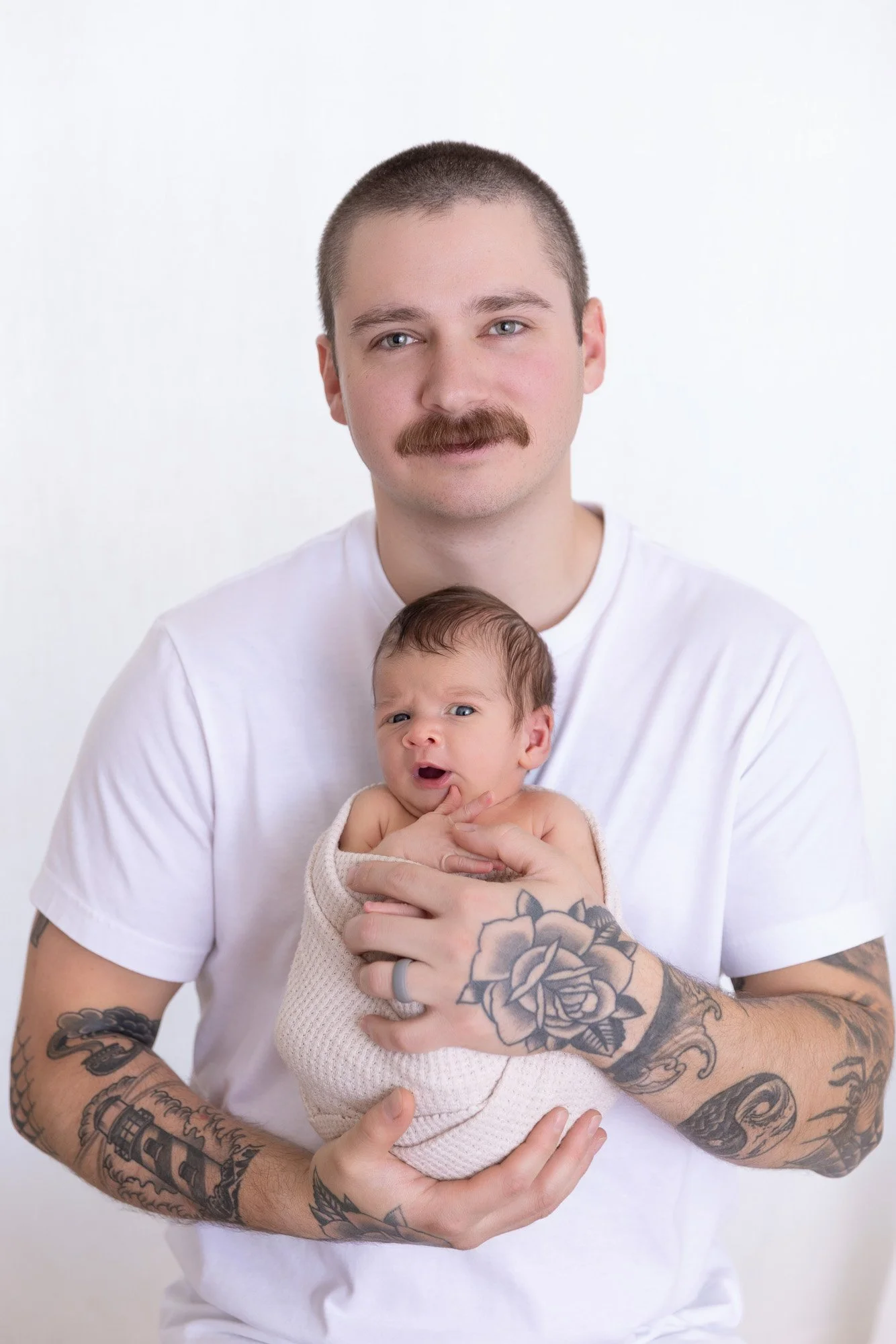 Photo of a father holding his newborn baby. Father has dark hair and blue eyes, with a mustache, and tattoos on the arms.  The baby has eyes open and is yawning.