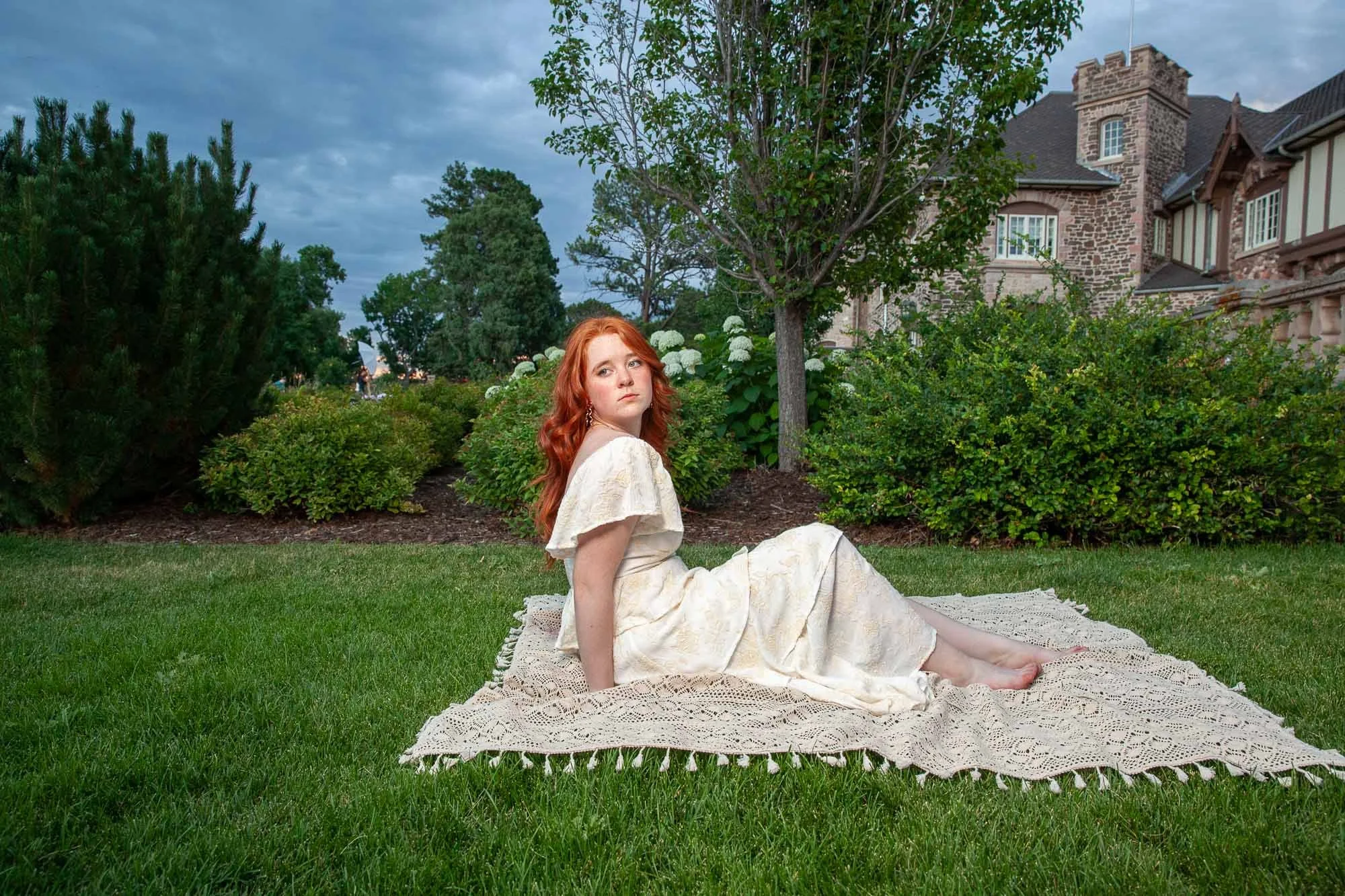 A young woman with red hair in a cream-colored dress sitting on a blanket in a garden with bushes, trees, and a stone house in the background.