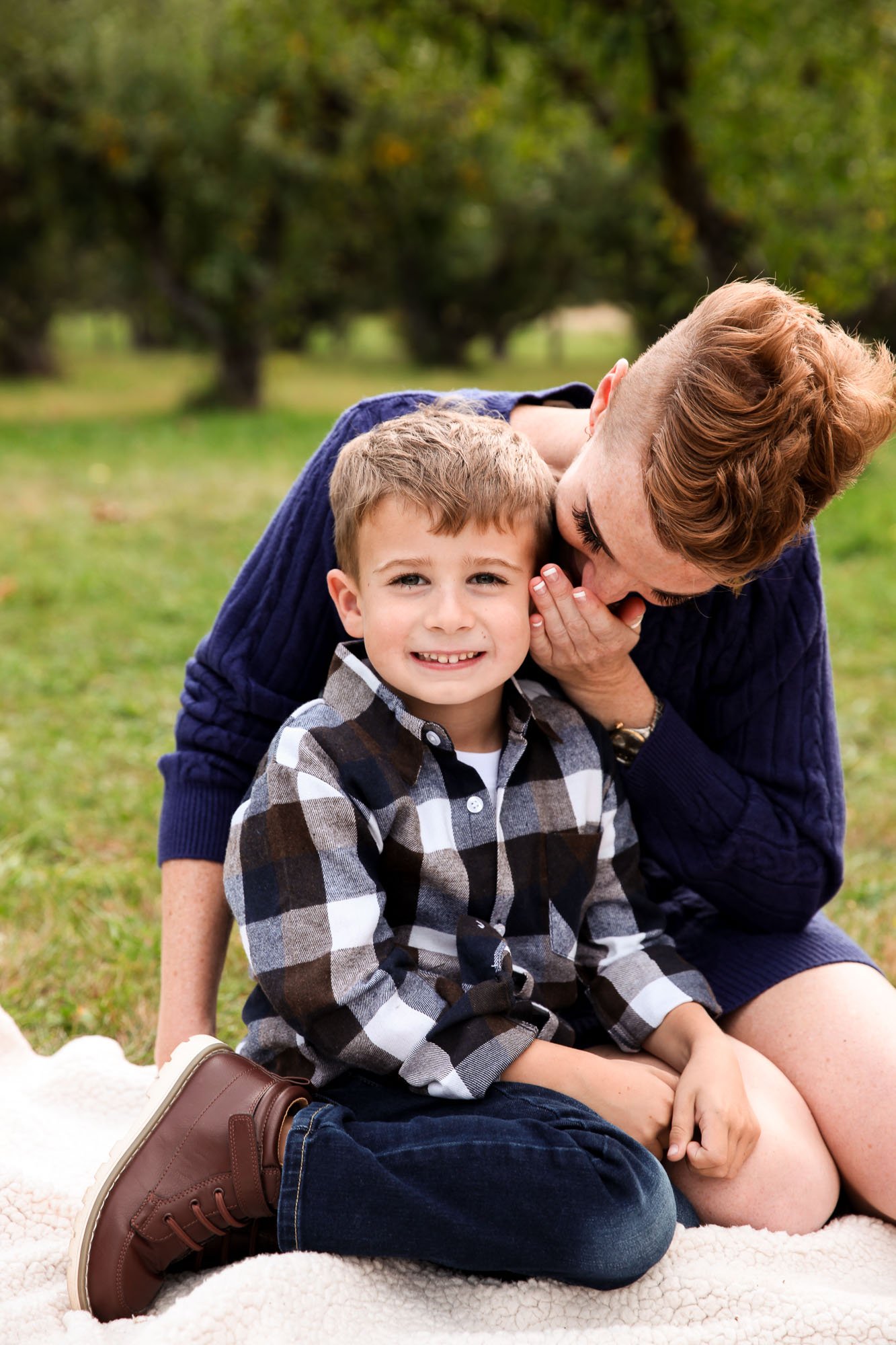 A child in a plaid shirt and brown boots sits on a blanket in a park, with an adult in a blue sweater whispering in their ear.
