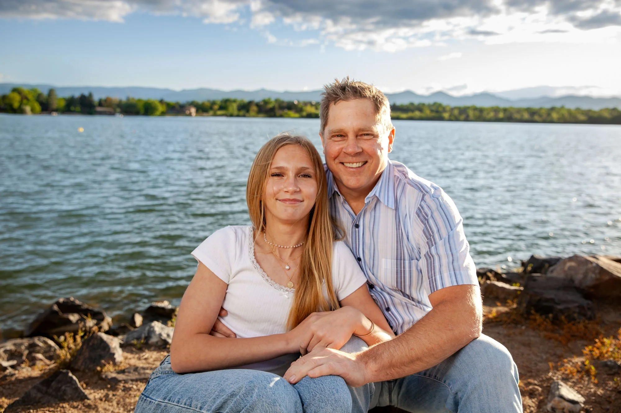 A smiling man and girl sitting on rocks by a lake during sunset, with mountains in the background.