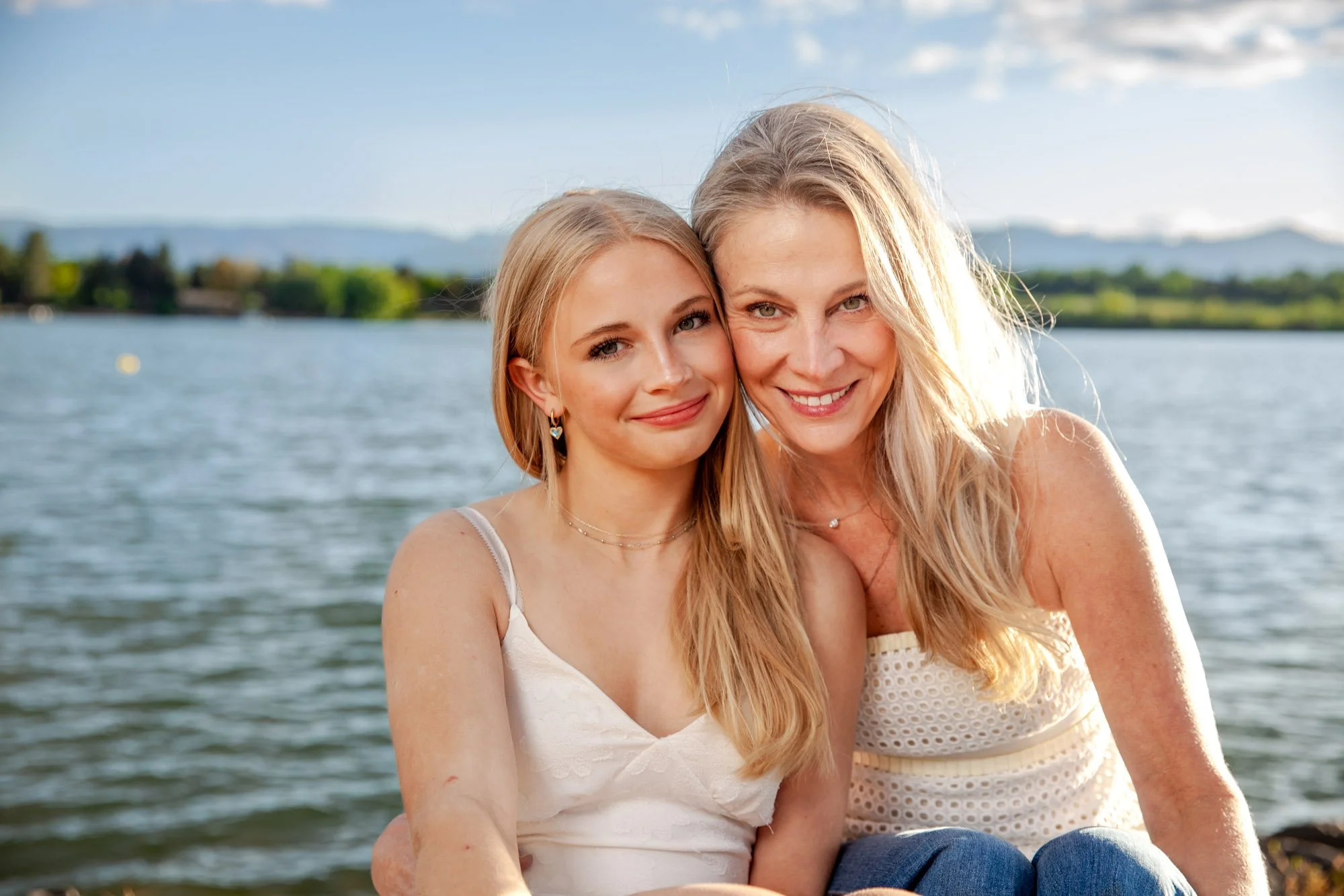 A woman and a teenage girl sitting by a lake, smiling at the camera on a sunny day.