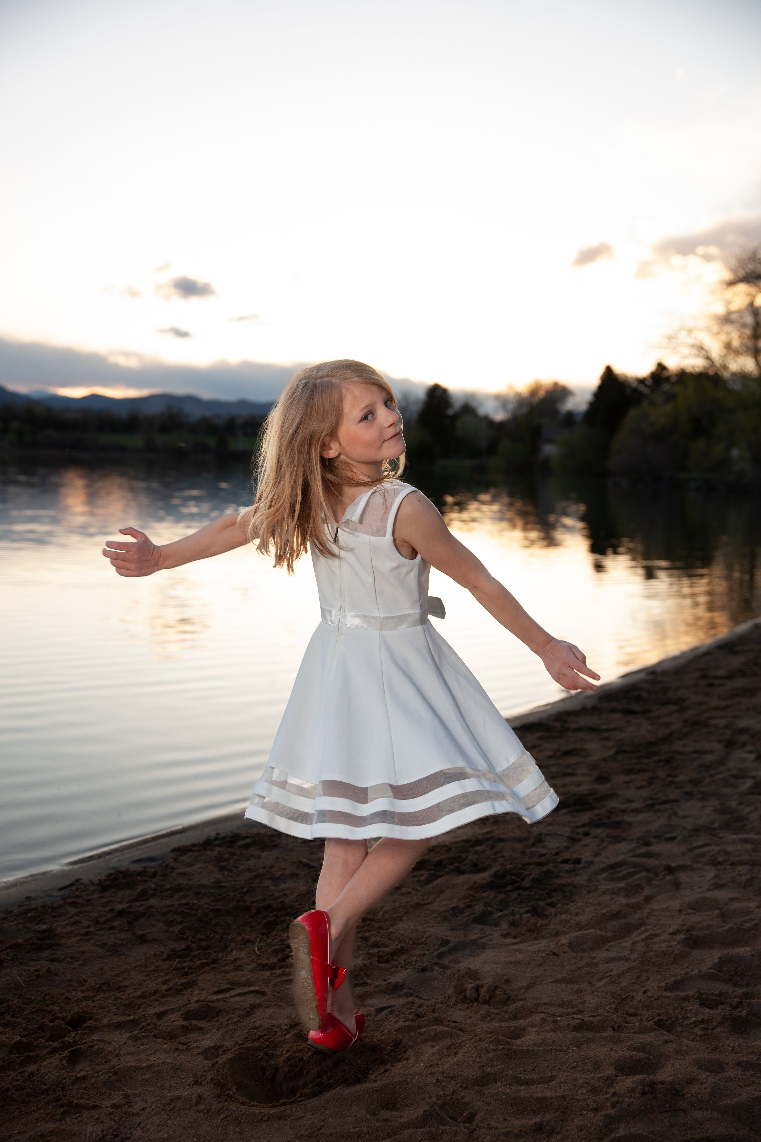 Young girl in a white dress and red shoes dancing on a sandy lakeshore at sunset.