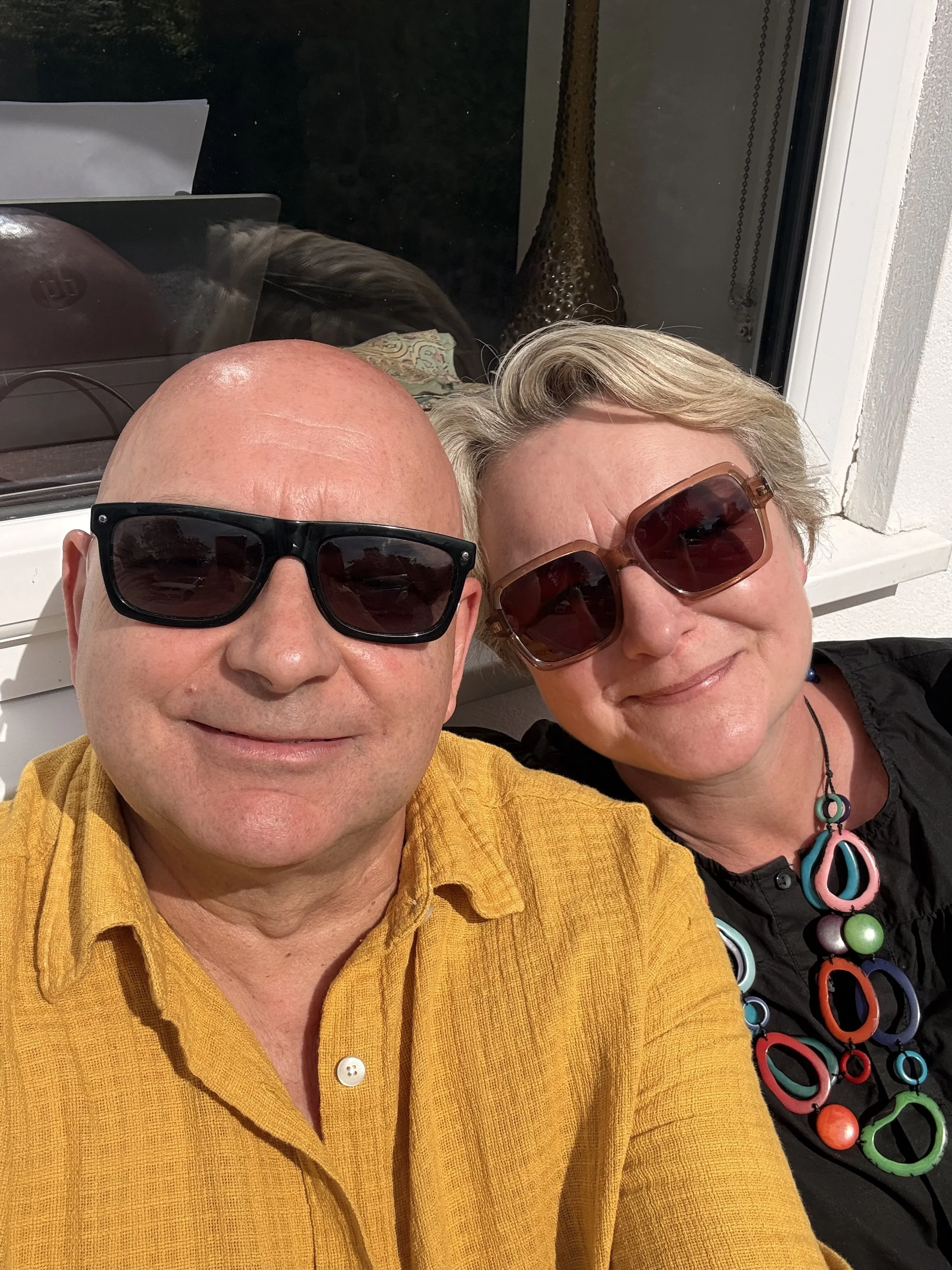 A close-up of a man and woman wearing sunglasses, smiling, and taking a selfie together. The man is wearing a yellow shirt, and the woman is wearing a black top with colorful jewelry. They are Emily & Graham Walters who own Shot Stop Craft Services