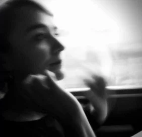Black and white photo of a smiling woman looking out of a moving vehicle window, with her hand resting on her chin.