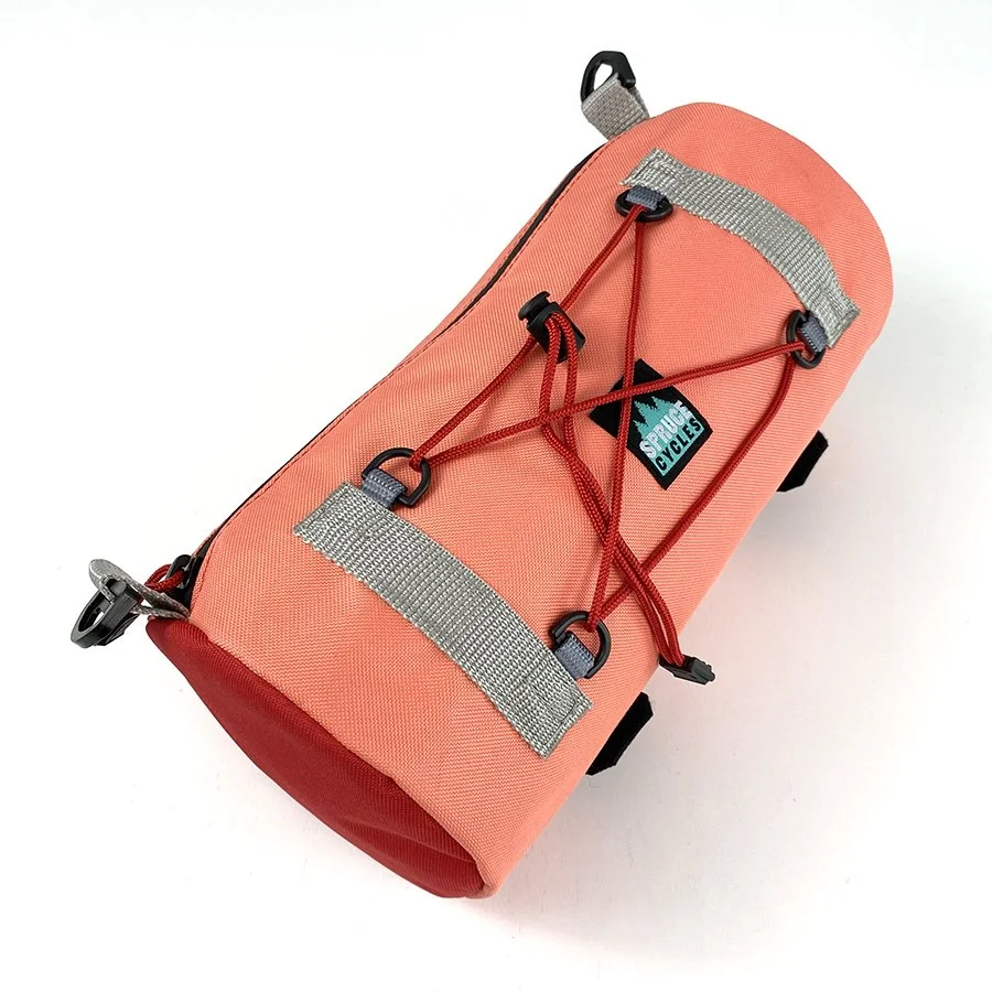 Coral-colored compact sports water bottle holder with gray straps and red bungee cords, branded with a Spruce Cyles logo patch.