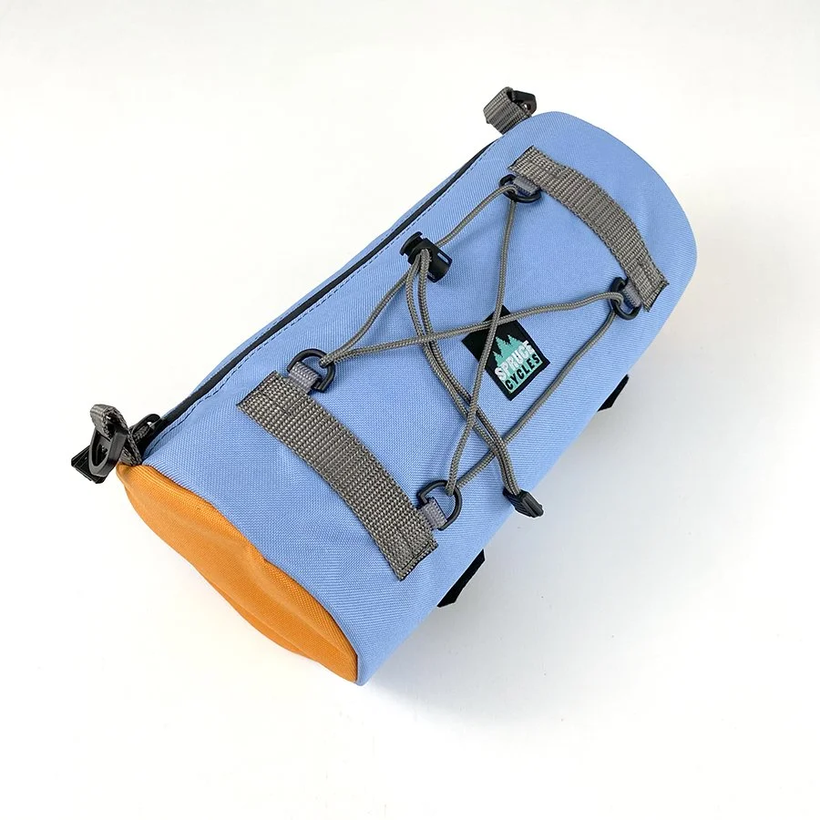 Light blue cylindrical outdoor gear bag with gray straps, orange bottom section, black clips, and a gray elastic cord with black hooks across the top, with a logo that says "Spruce Cycles" on a black patch.