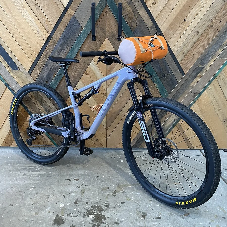A mountain bike with a light blue frame leaning against a wooden wall with geometric pattern, with an orange cargo bag attached to the handlebar.