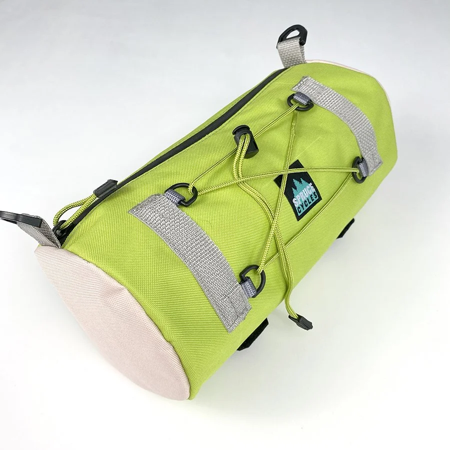 Green and pink sports duffel bag with exterior bungee cord, gray straps, and a logo that says 'Spruce Cycles' on the side.