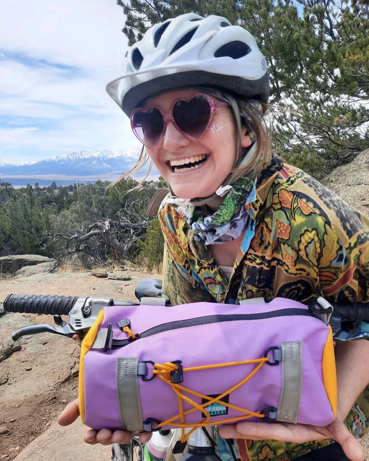 A smiling woman wearing a white bike helmet, heart-shaped sunglasses, and a colorful shirt, holding a purple and yellow bike bag with orange bungee cords on a mountain trail with trees, rocks, and snow-capped mountains in the background.