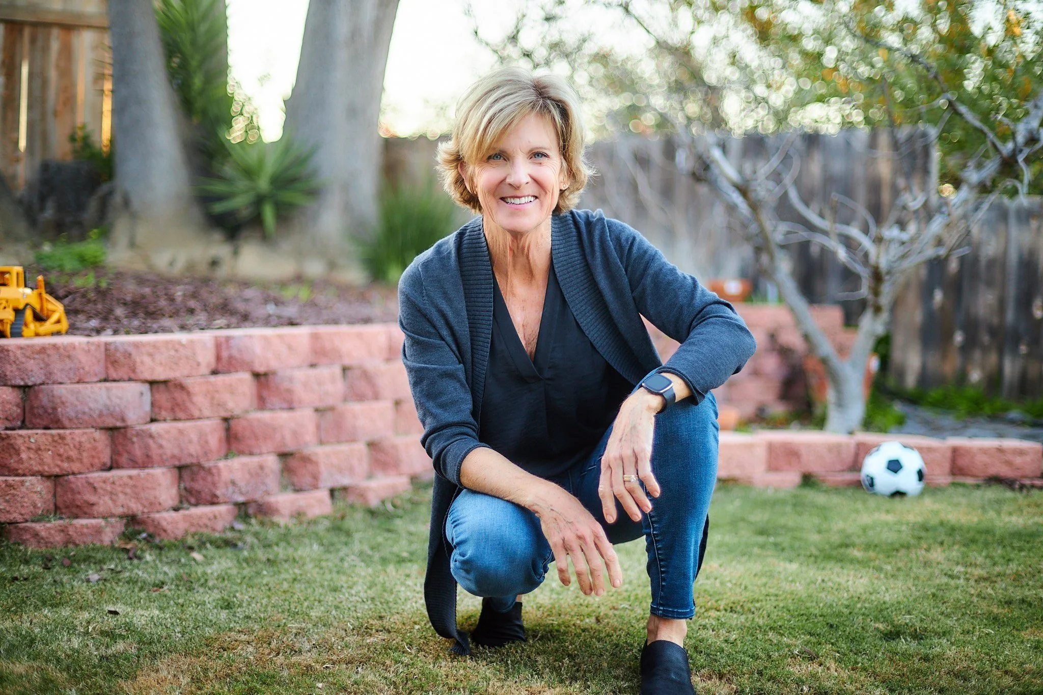 Person squatting in a backyard with a brick wall, soccer ball, and trees
