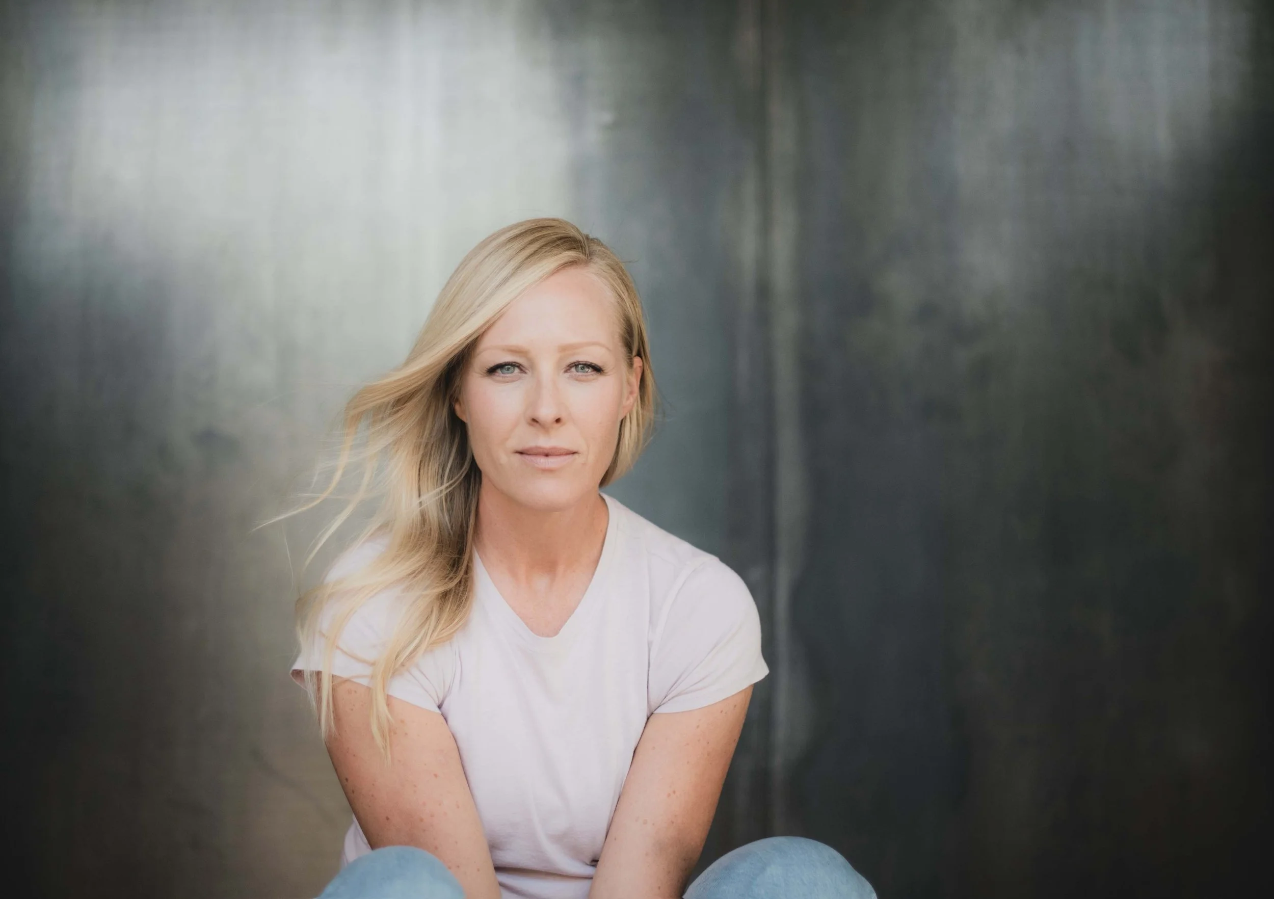 A woman with long blonde hair and blue eyes, wearing a white t-shirt, sitting against a metallic background.