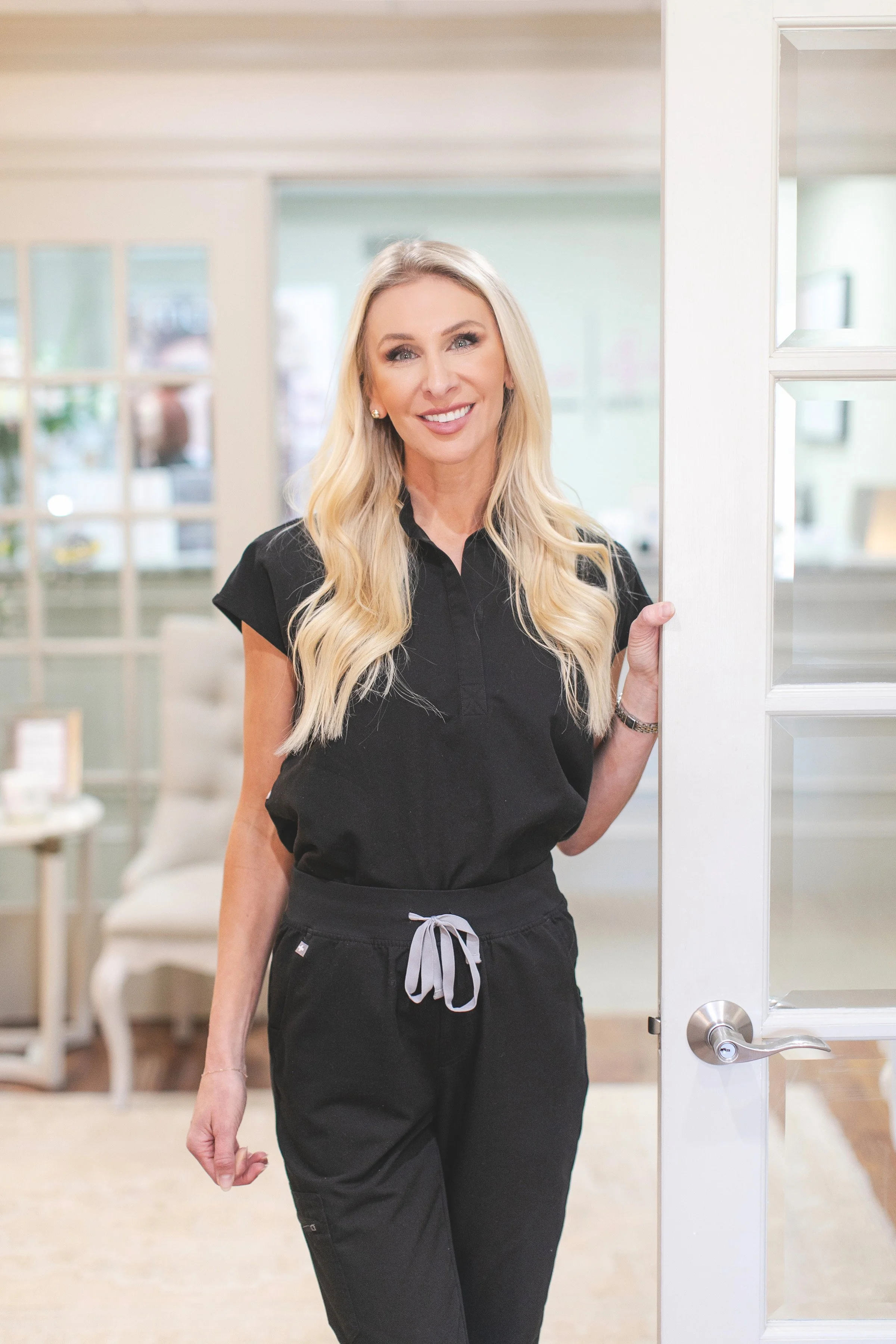 A woman with long blonde hair wearing a white blouse and maroon skirt standing in a room, smiling and holding a beauty tool in her right hand.