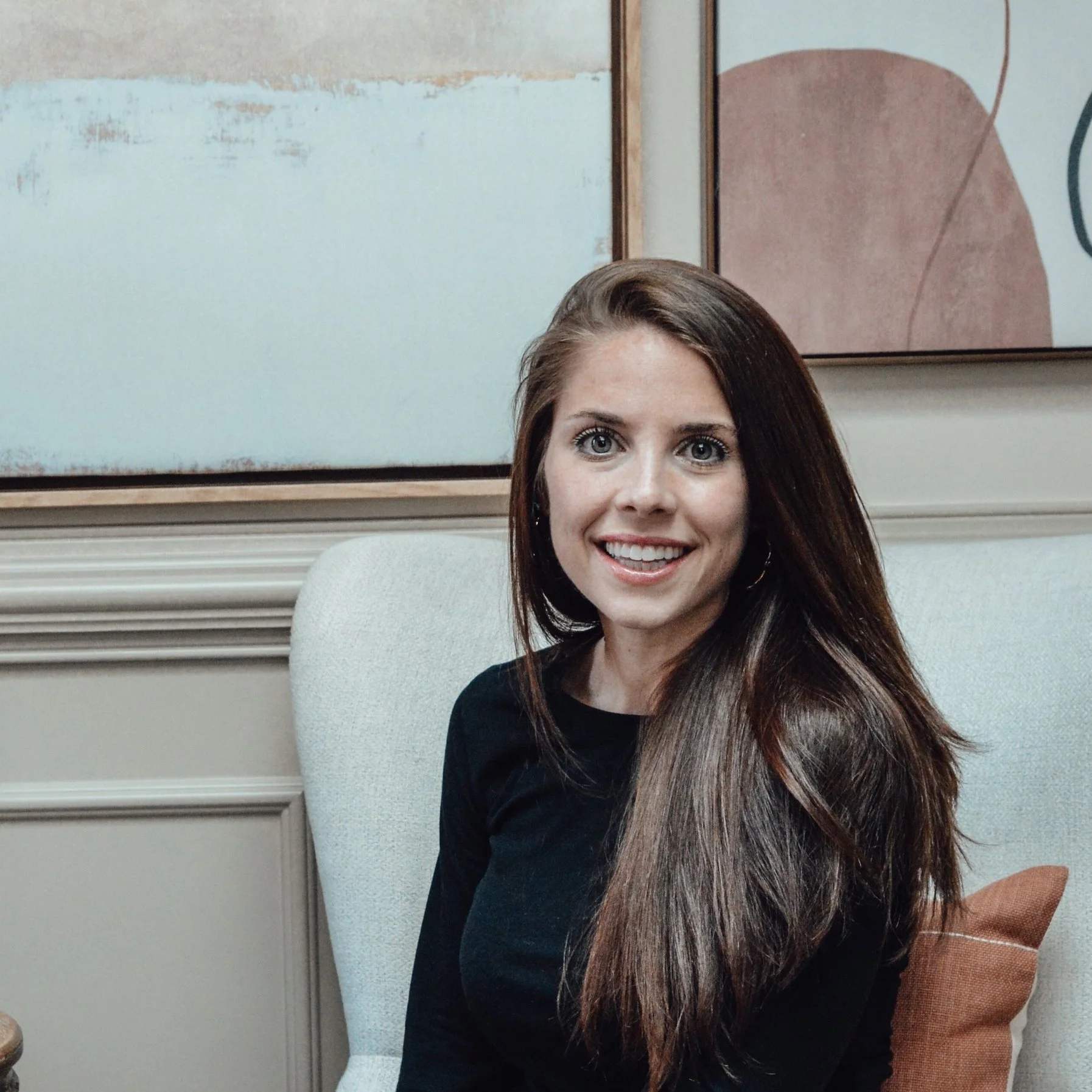 A smiling woman with long brown hair, wearing a black top, sitting on a beige armchair in a room with modern art on the wall.